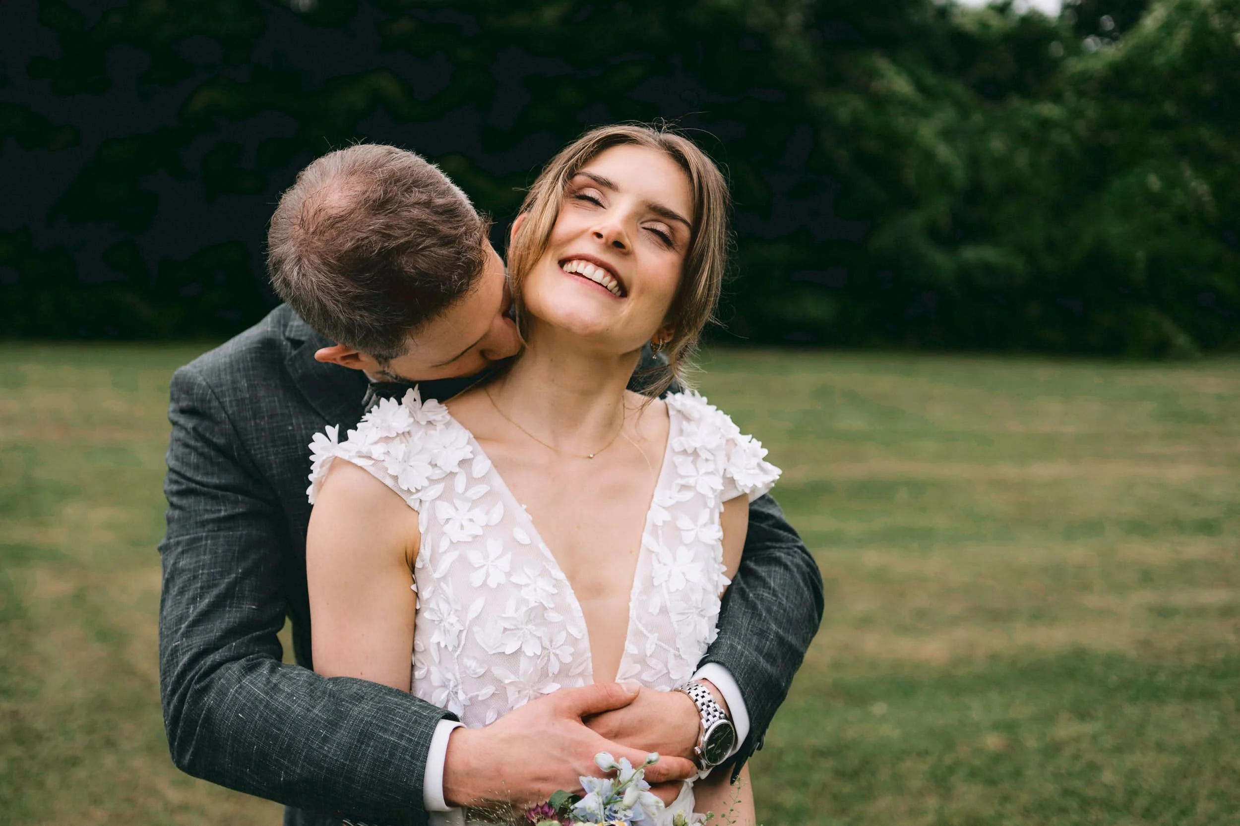 Ein glückliches Paar bei einer Hochzeit im Freien, der Mann umarmt die Frau von hinten und küsst sie auf die Wange, sie lächelt mit geschlossenen Augen, im Hintergrund Bäume und Wiesen.