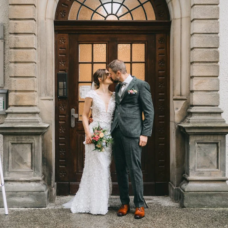 Ein Brautpaar steht vor einer alten Holztür, küsst sich und trägt formelle Hochzeitskleidung, die Braut hält einen Blumenstrauß.