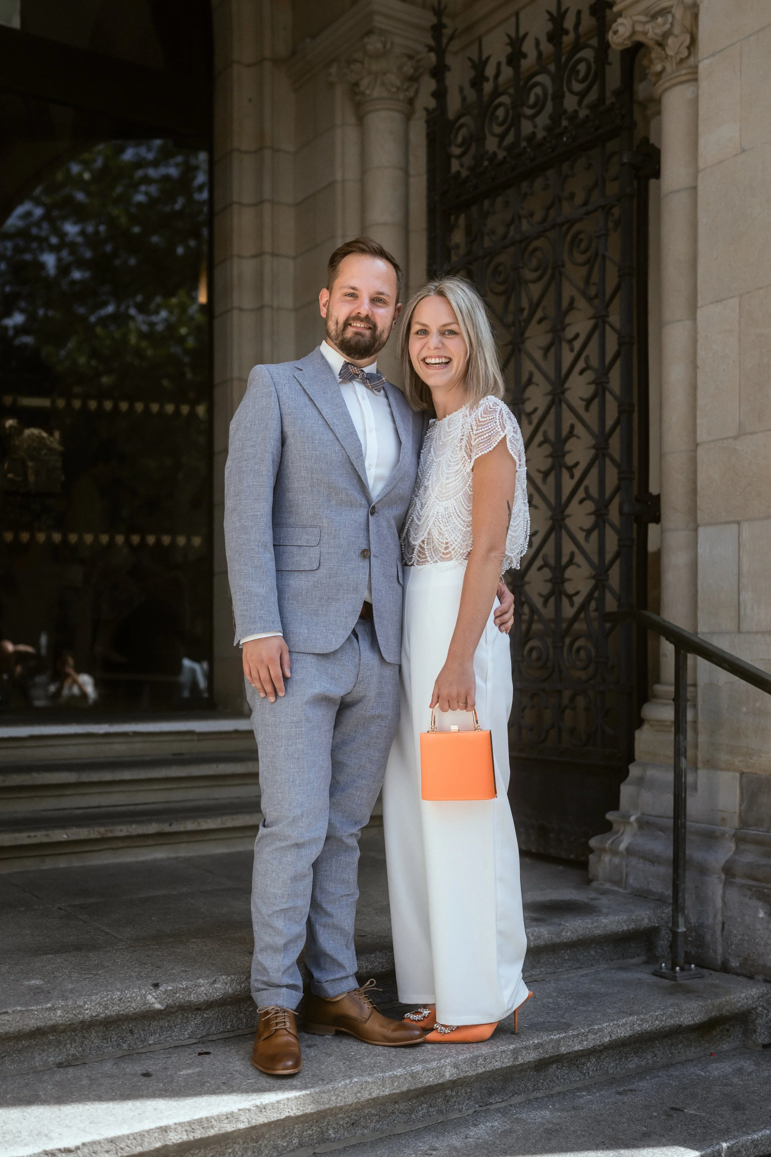 Ein Mann und eine Frau posieren an einer Treppe vor einem historischen Gebäude mit schmiedeeisernem Tor. Der Mann trägt einen grauen Anzug mit Krawatte, die Frau trägt ein weißes Kleid mit Spitze und hält eine orange Tasche. Beide lächeln in die Kame