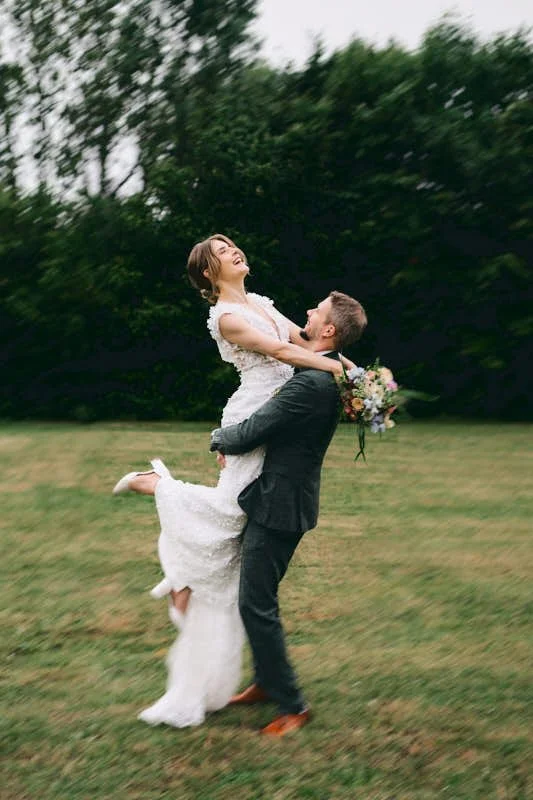Ein braunhaariger Mann in einem dunklen Anzug hebt eine lachende Braut mit kurzen braunen Haaren in einem weißen Hochzeitskleid. Sie hält einen Blumenstrauß in der Hand, während sie auf einer grünen Wiese vor dunklen Bäumen steht.