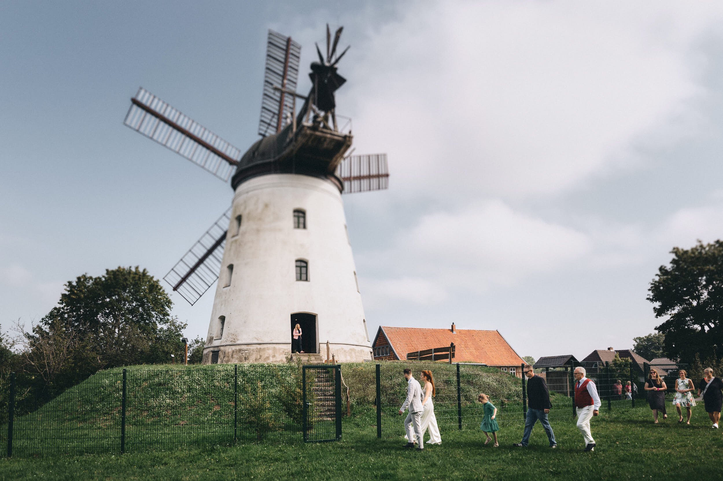 Eine weiße Windmühle mit schwarzen Fenstern und roten Flügeln steht in einer grünen Landschaft. Im Vordergrund laufen mehrere Menschen, darunter ein Paar in formeller Kleidung, auf einem grasbewachsenen Hügel, umgeben von einem Zaun und Bäumen.