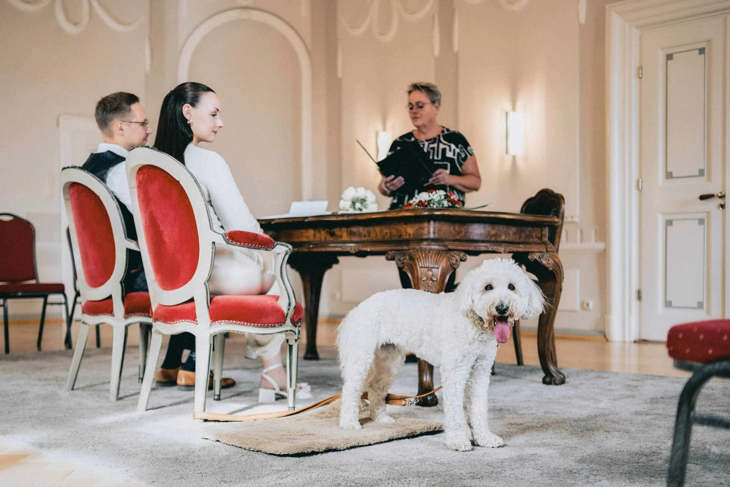 Ein Hund liegt auf einem Teppich im Vordergrund, während drei Menschen, zwei Frauen und ein Mann, in festlicher Kleidung an einem Tisch sitzen. Eine Frau liest aus einem Buch vor, während die anderen zuhören. Der Raum ist elegant eingerichtet mit rot