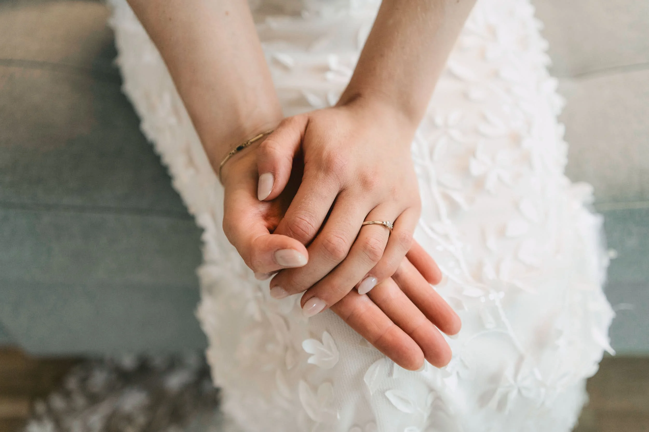 Nahaufnahme von zwei Händen, die eine goldene Ehering tragen, auf einem weißen, bestickten Hochzeitskleid.