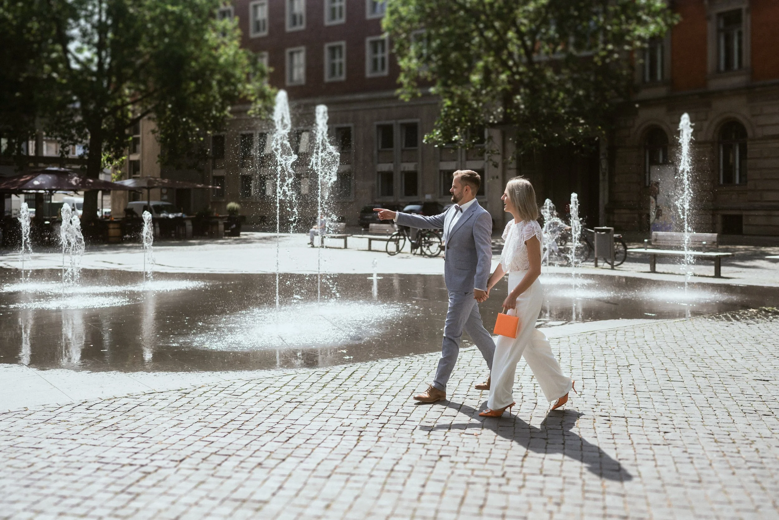 Ein Paar in formeller Kleidung spaziert Hand in Hand an einem Springbrunnen in einer Stadt. Der Mann trägt einen grauen Anzug, die Frau ein weißes Kleid, und beide haben ein Lächeln im Gesicht.