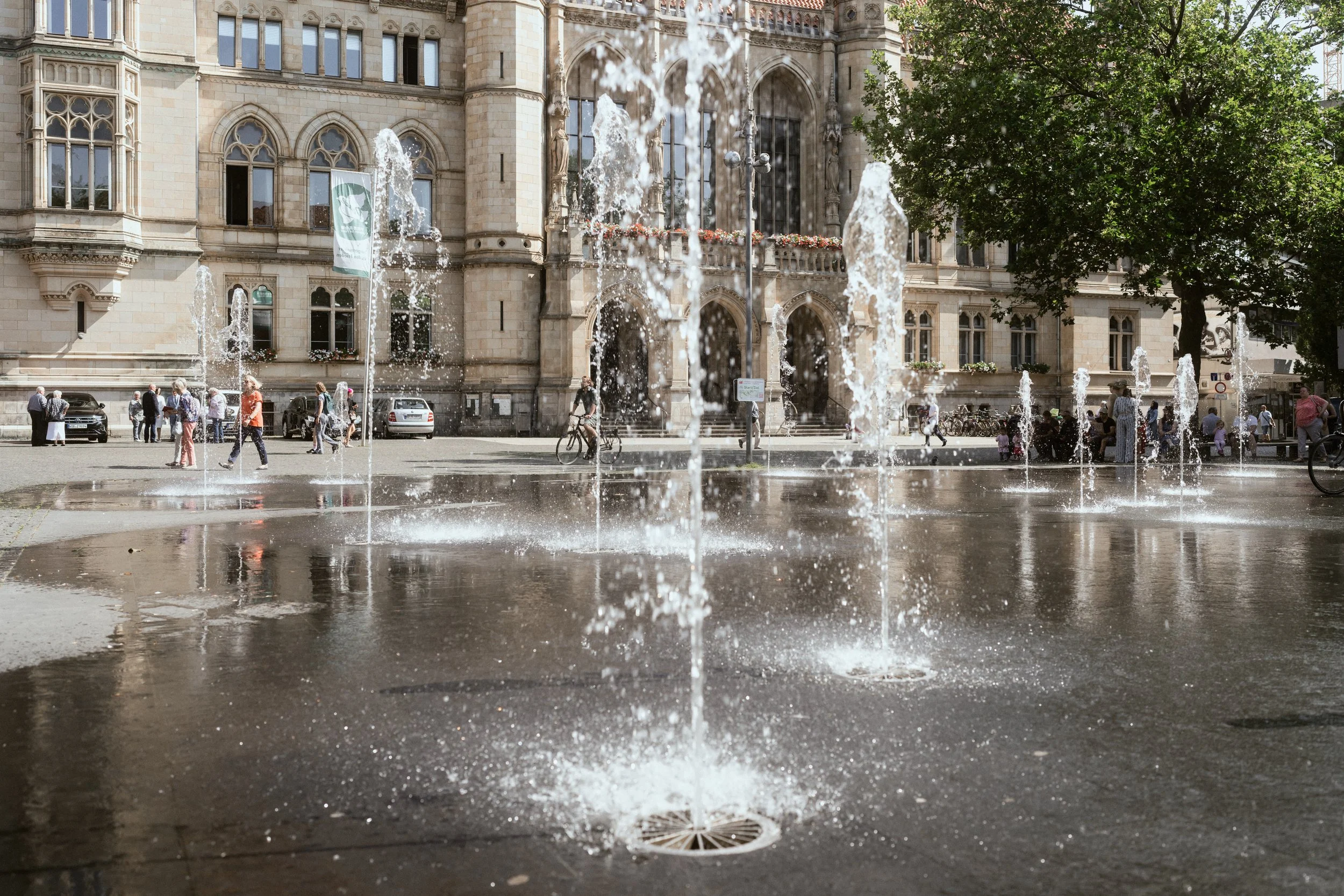 Blick auf einen Brunnen in einer Stadt mit einem historischen Gebäude im Hintergrund, Menschen laufen, Fahrradfahrer fährt durch den Brunnenbereich, Bäume auf der Seite, Wasser spritzt aus den Brunnen.