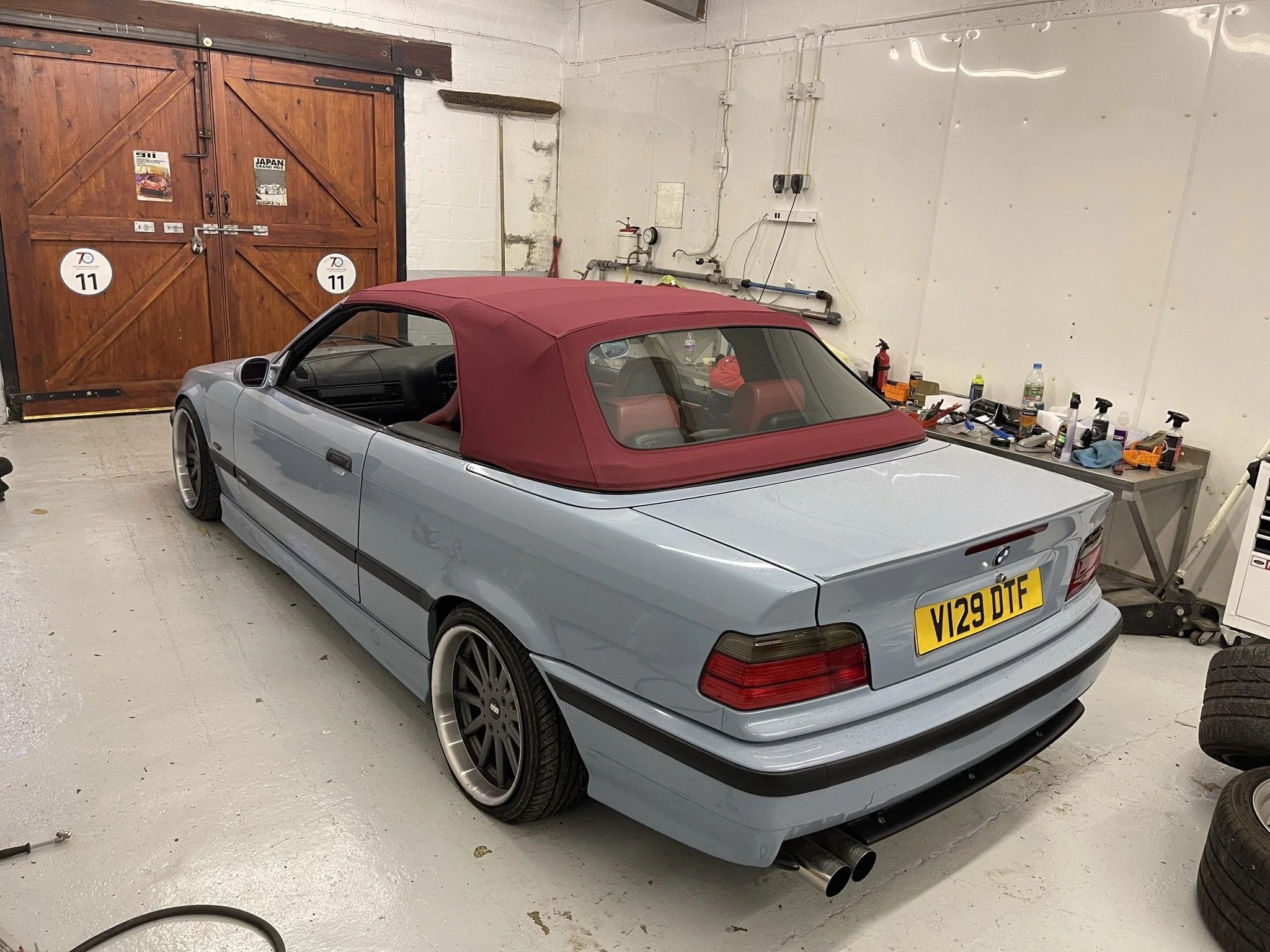 A light blue convertible car with a red soft top parked inside a garage. The car has a yellow UK license plate with the registration V129 DTF. The garage has a wooden door, a work table with tools and bottles, and some tires stacked on the side.