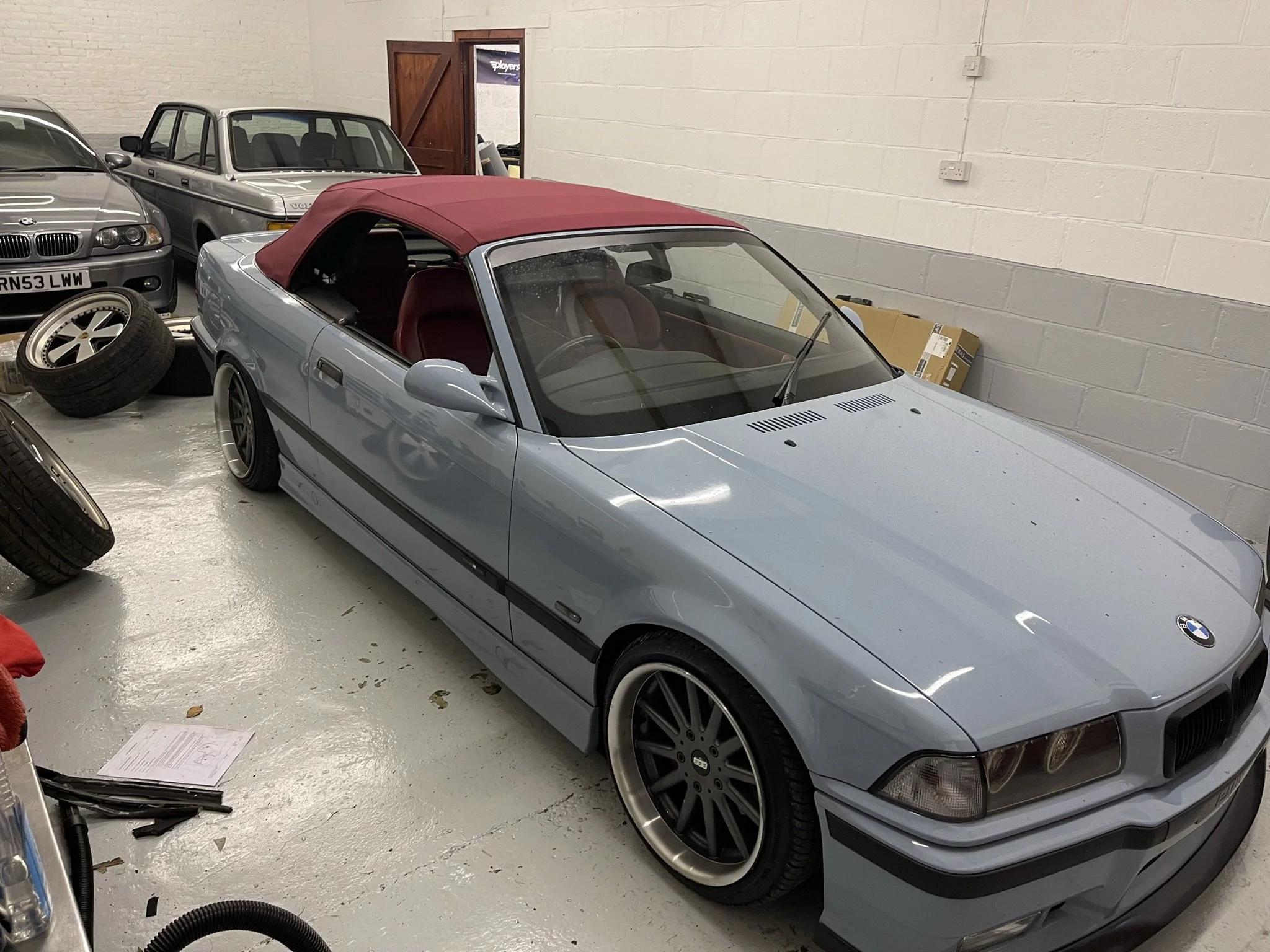 Light blue BMW convertible with a red soft top parked inside a garage, surrounded by other classic cars and spare tires.