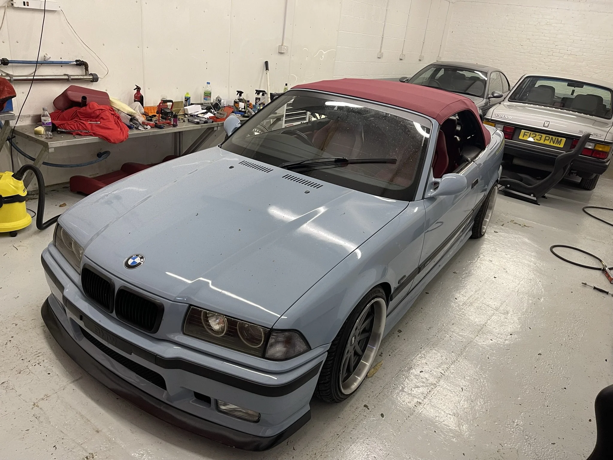 Light blue BMW convertible with a maroon soft top parked in a garage, surrounded by car tools and two other vehicles, one of which is a silver Volvo with a yellow license plate.