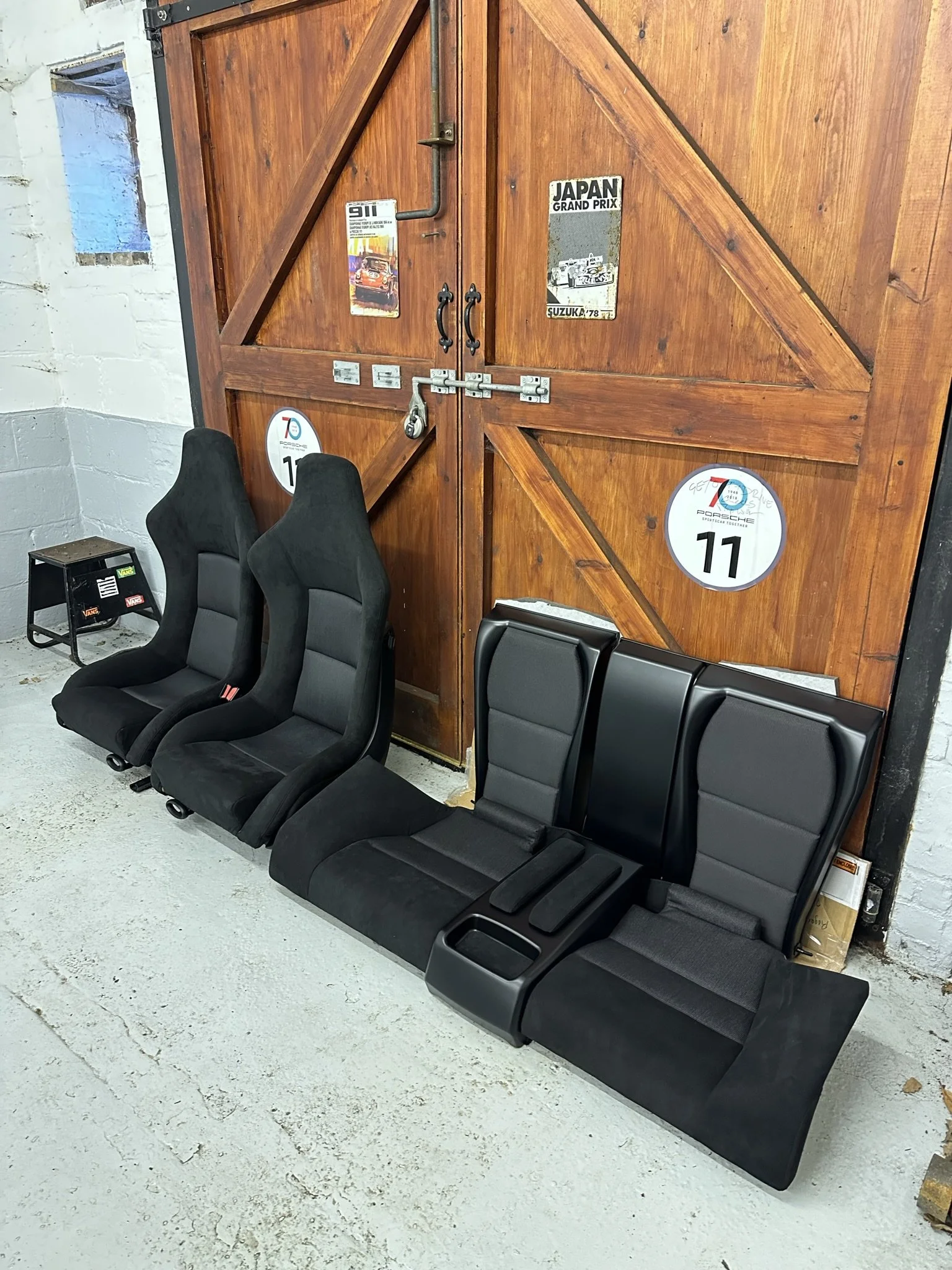 Set of four black car interior seats placed against a large wooden barn door with vintage car posters and signs.