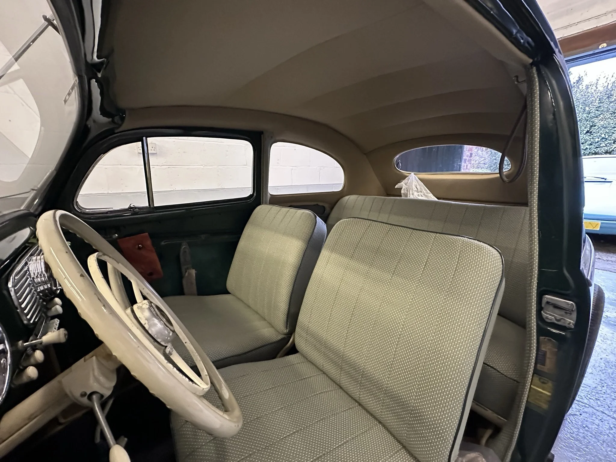 Interior of a vintage car showing a cream-colored steering wheel, dashboard, and beige upholstered seats. The car has a rounded dashboard and large windows, with a small rear window visible.