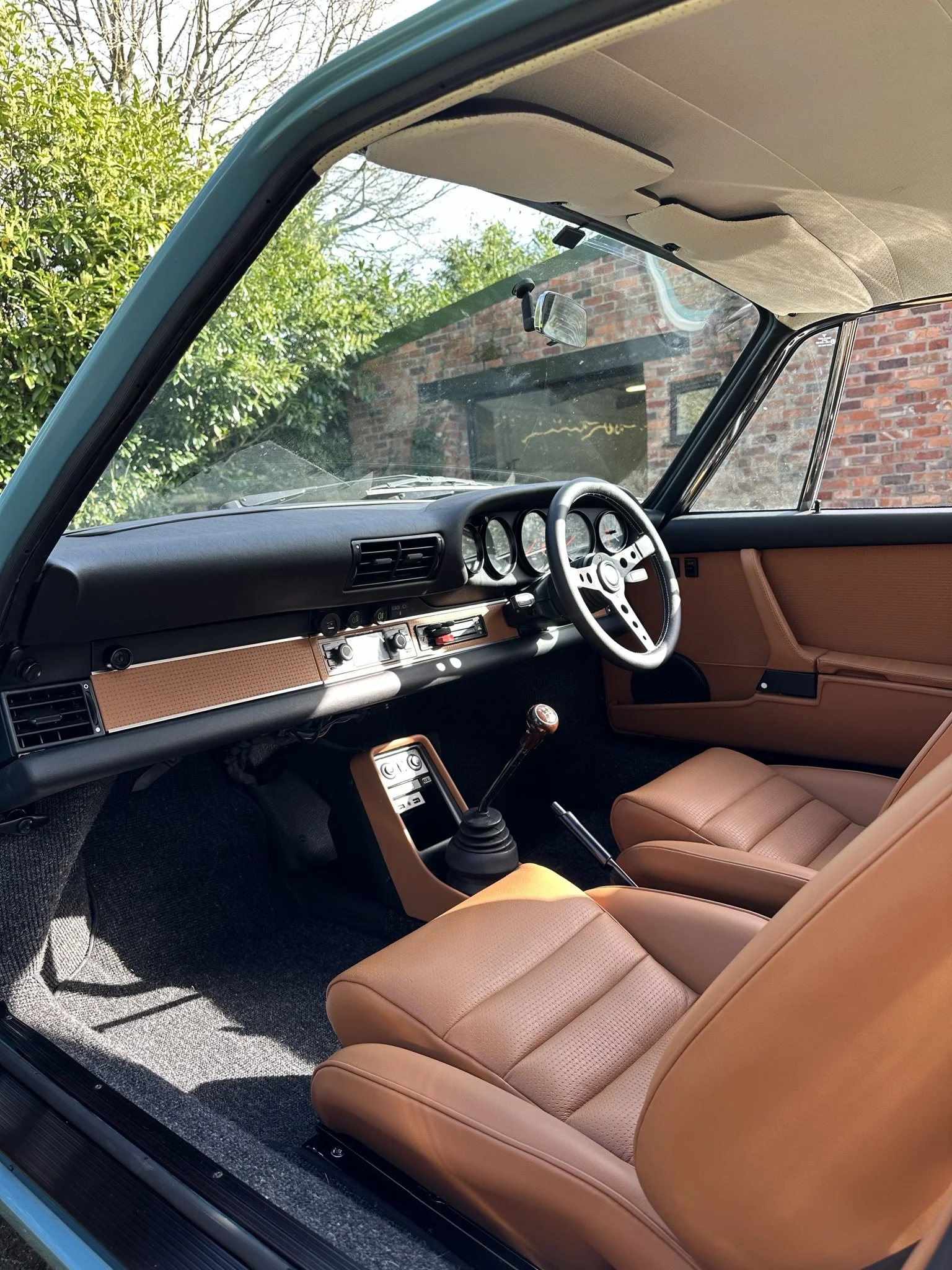 Interior of a vintage car with tan seats, black dashboard, and a manual gear stick, viewed from the open driver's side door.