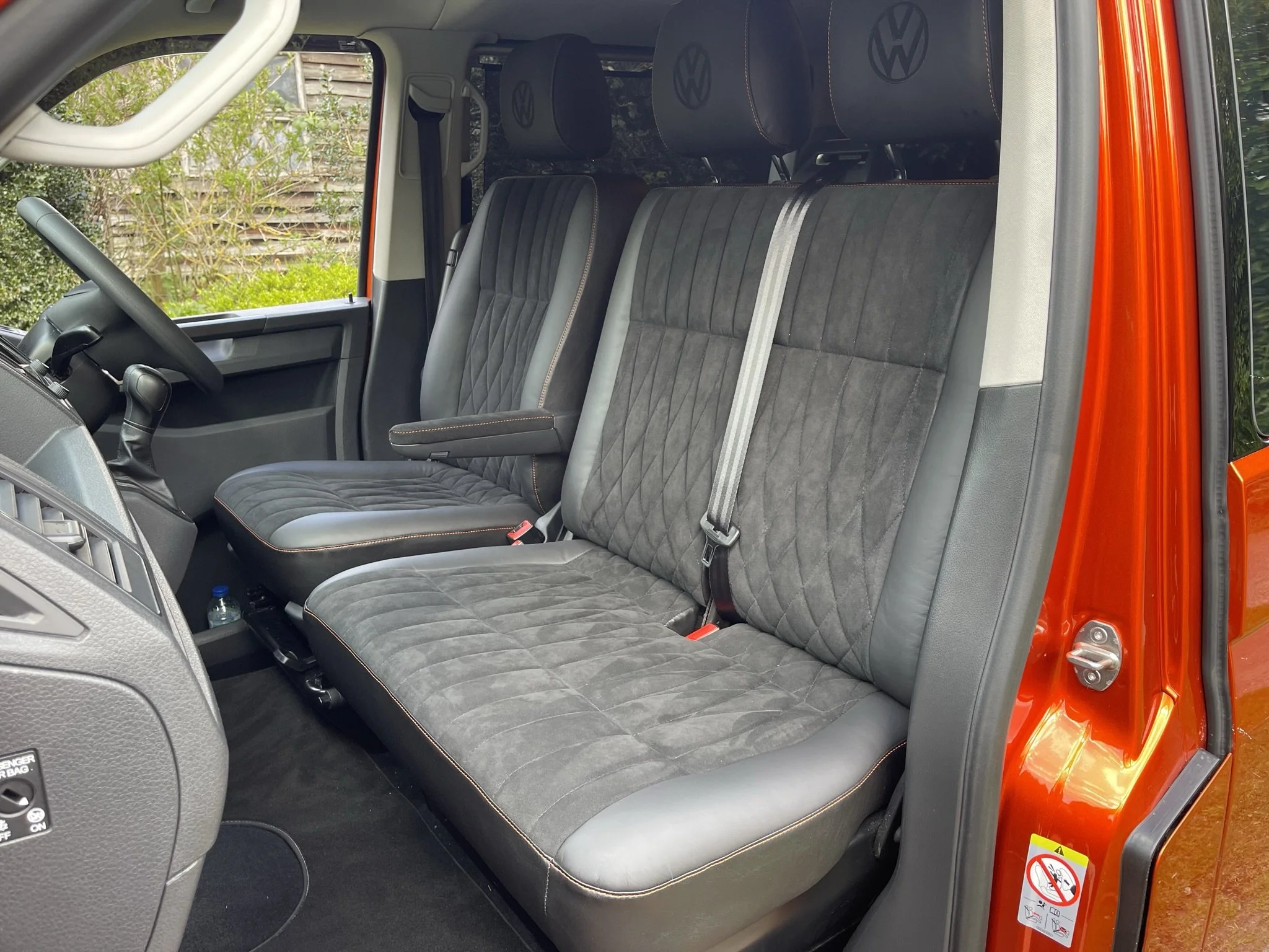 The interior of a Volkswagen vehicle showing the front passenger seat and the rear bench seat, both with black upholstery and orange stitching, in a vehicle with a red exterior.