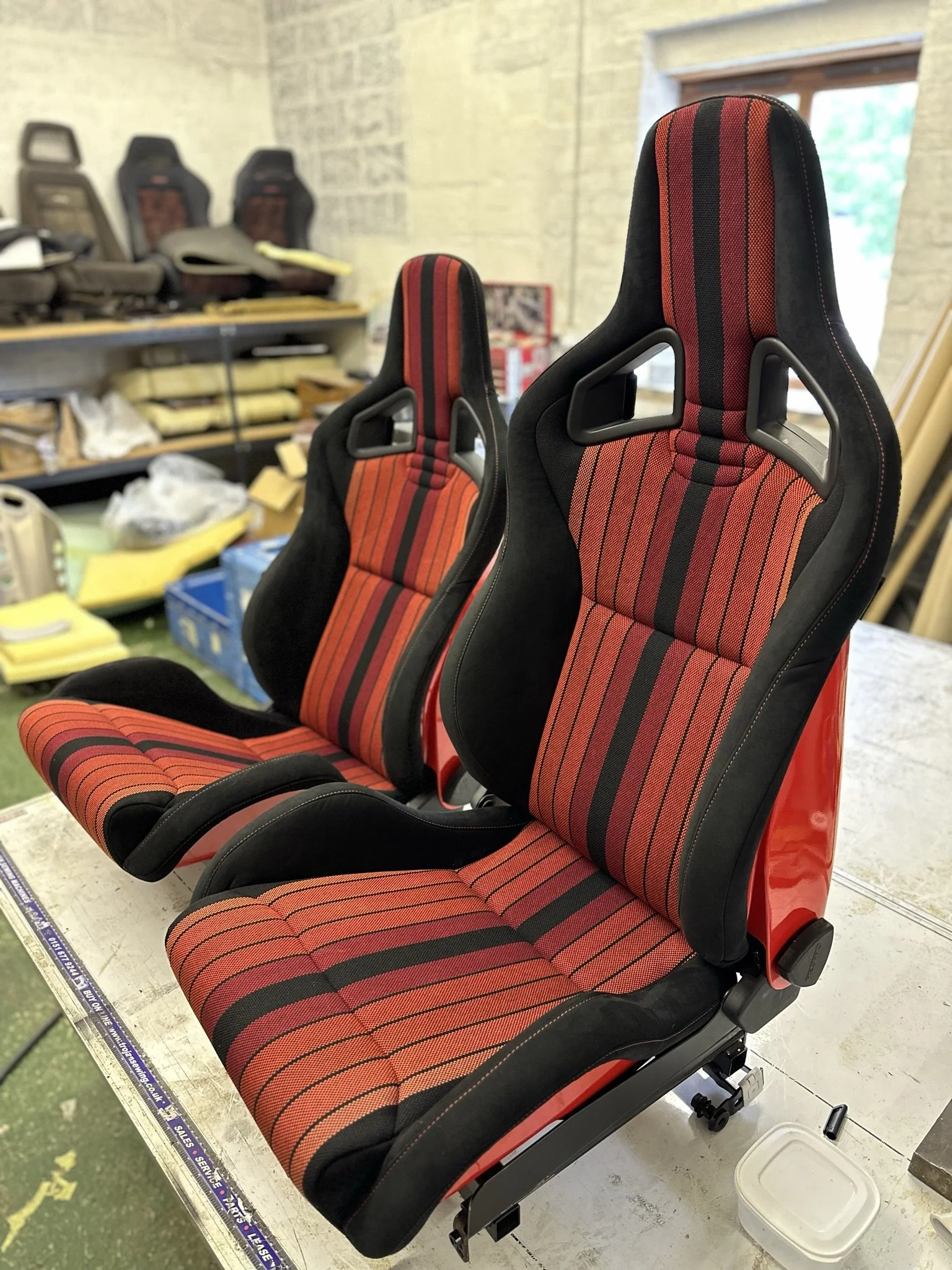 Two racing car seats with red and black striped fabric upholstery, placed on a workshop table.