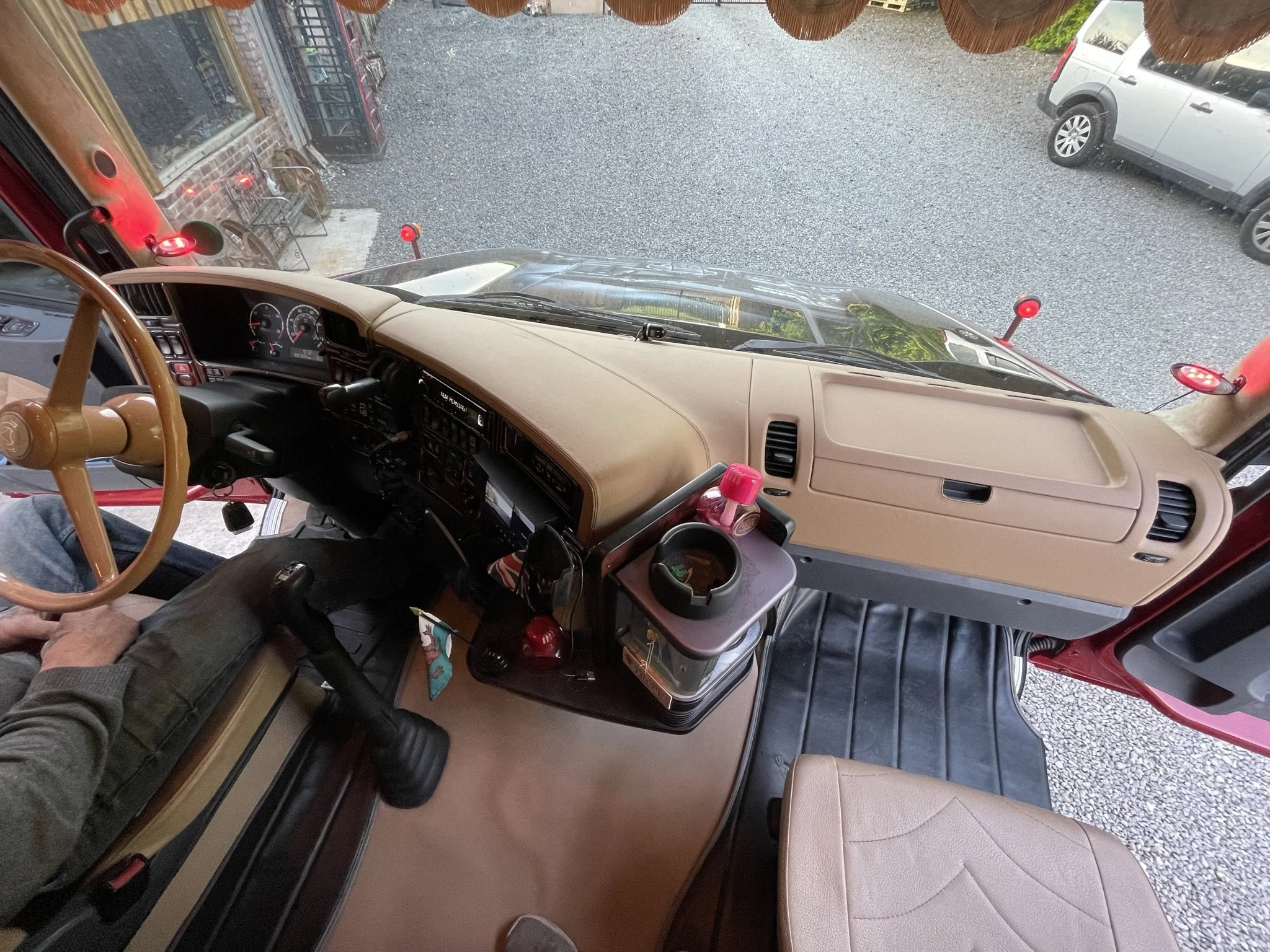 Inside the cab of a semi-truck showing the dashboard, steering wheel, and passenger seat, with parking lot visible through the windshield.