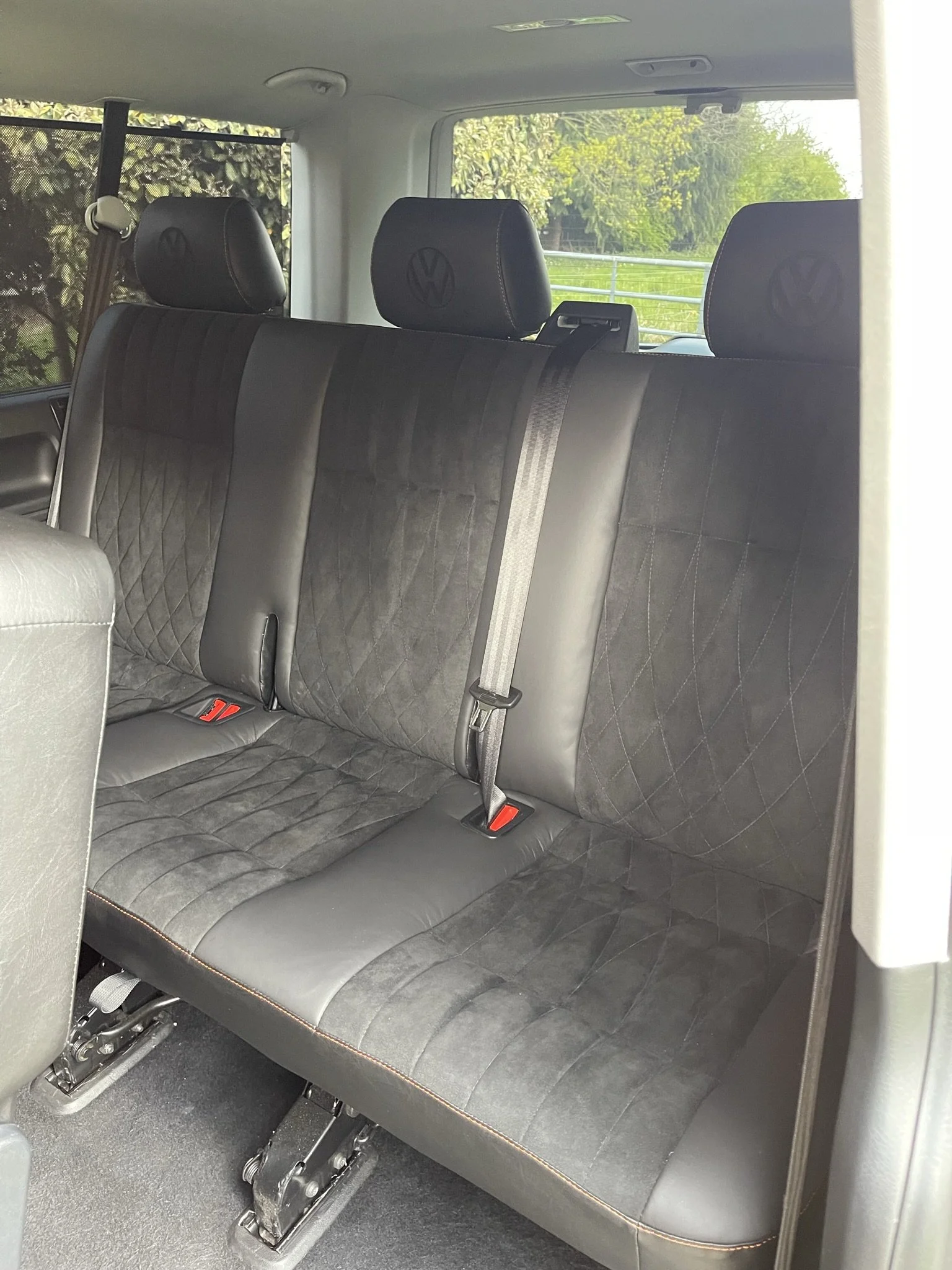 Interior of a vehicle showing a dark-colored, three-row rear bench seat with headrests, seat belts, and window view of greenery outside.