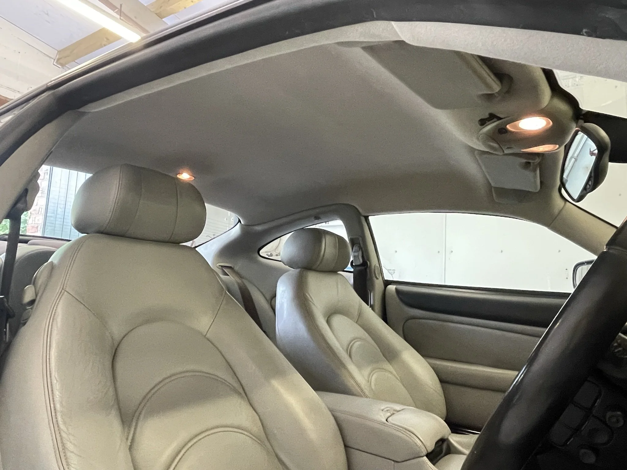 The interior of a car showing beige leather seats, overhead interior lights, and a rearview mirror.