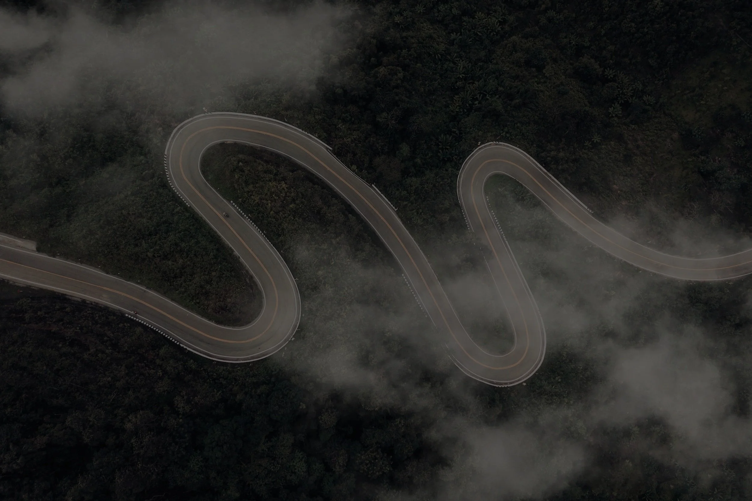 An aerial view of a winding mountain road with sharp curves, surrounded by dense forest and low-hanging clouds.