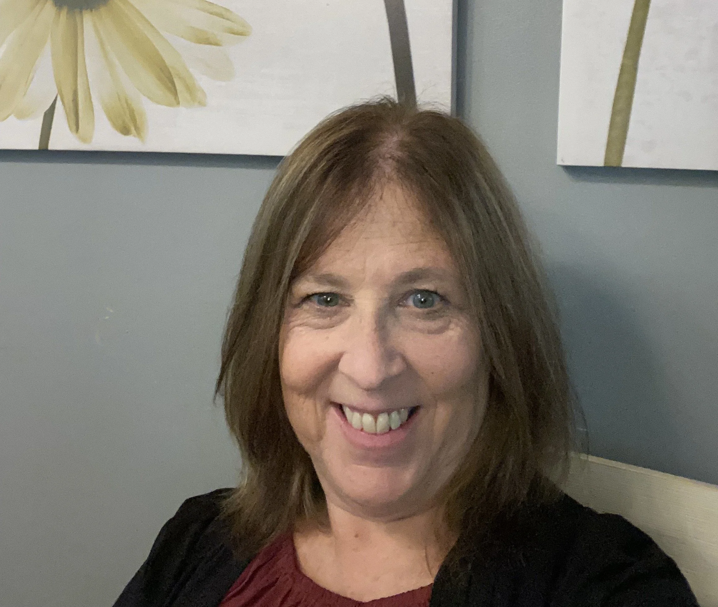 Smiling woman with shoulder-length brown hair and blue eyes, wearing a black jacket and a maroon top, is sitting in a room with blue walls and floral artwork in the background.