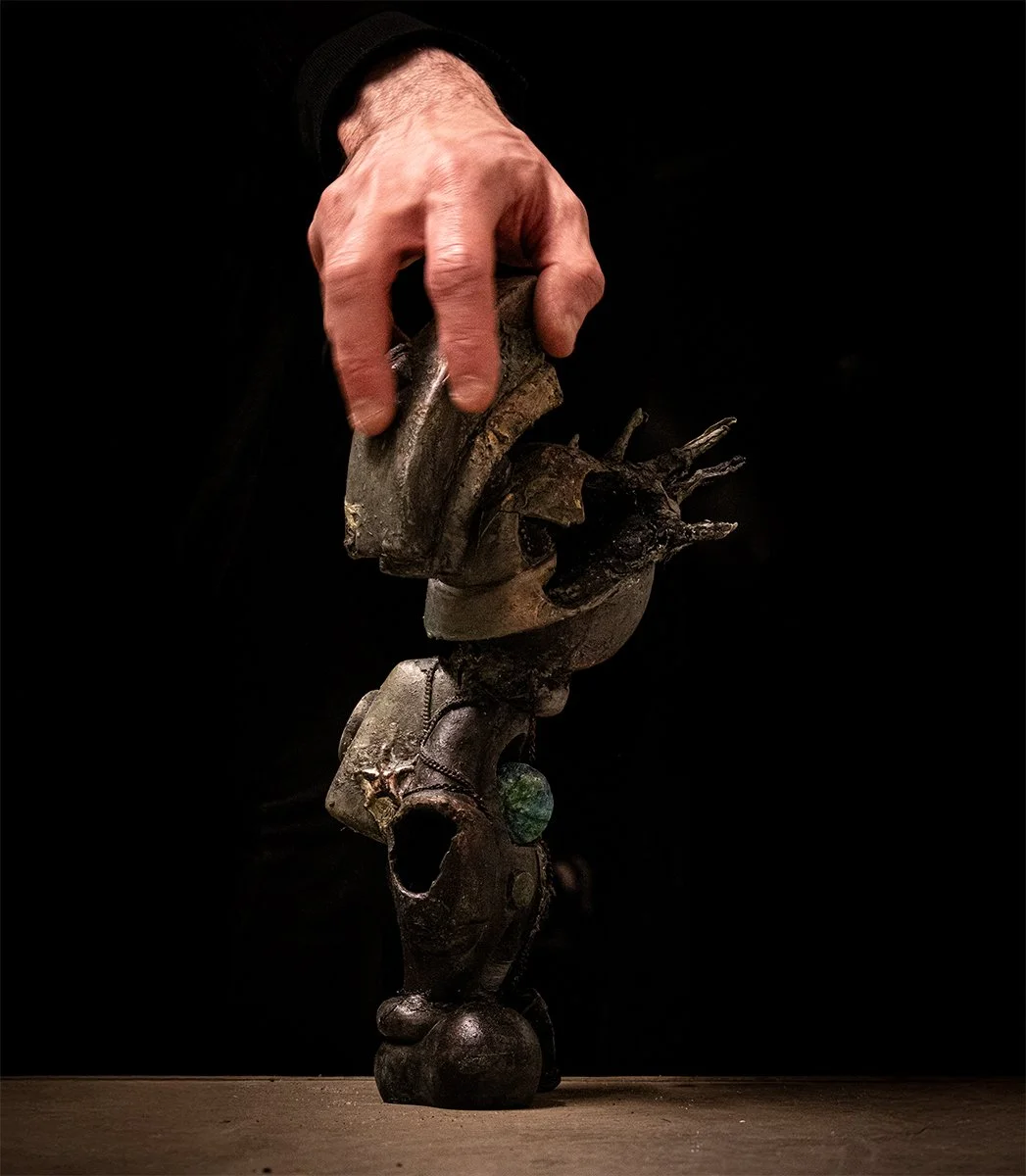 A person with a hand in a black sleeve is arranging or holding a dark, abstract sculpture made of various metallic and wooden materials against a black background.