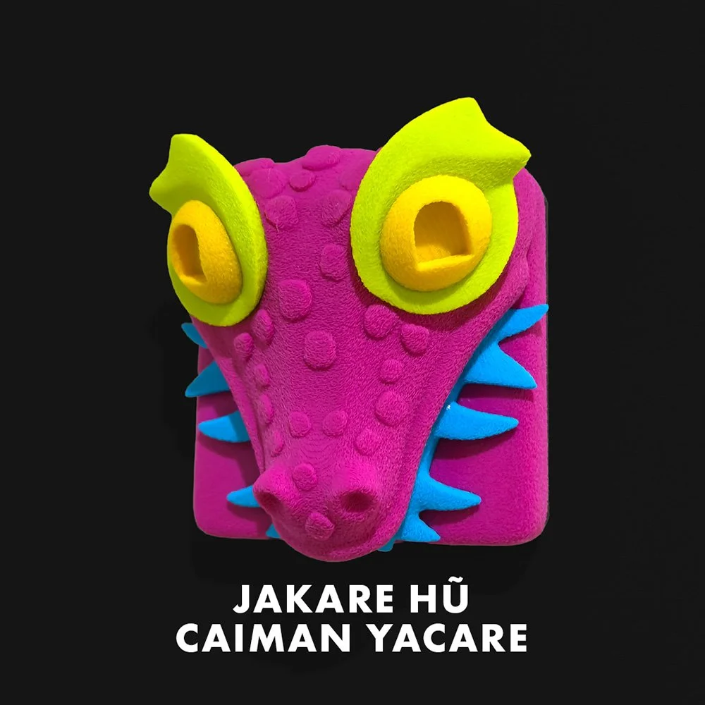Colorful, plush monster toy with pink face, yellow eyes, blue spikes, and black background. Text below reads "JAKARE HÜ CAIMAN YACARE."