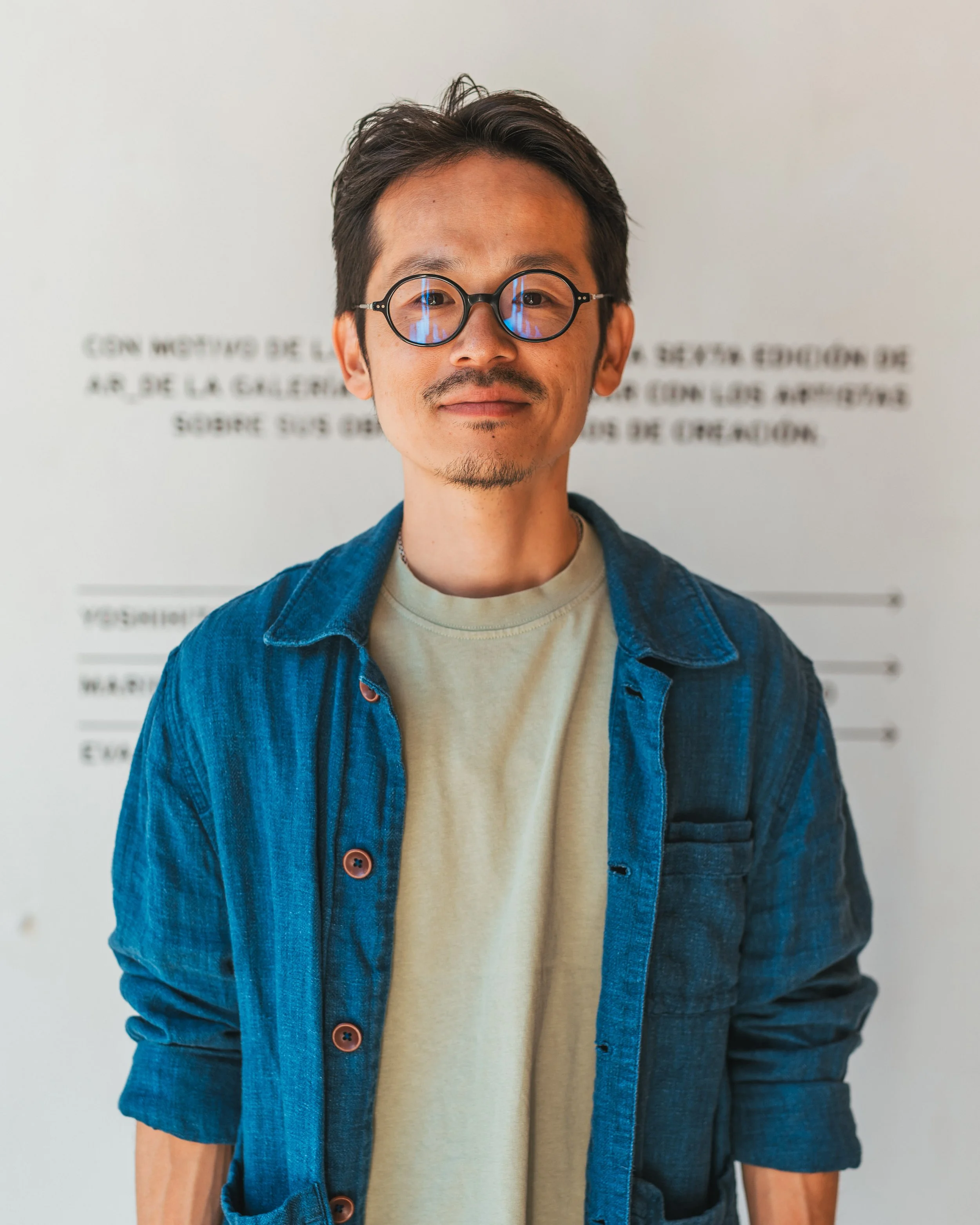 A man with short dark hair, glasses, and a slight beard, wearing a beige t-shirt and a blue denim jacket, standing in front of a white wall with blurry text.
