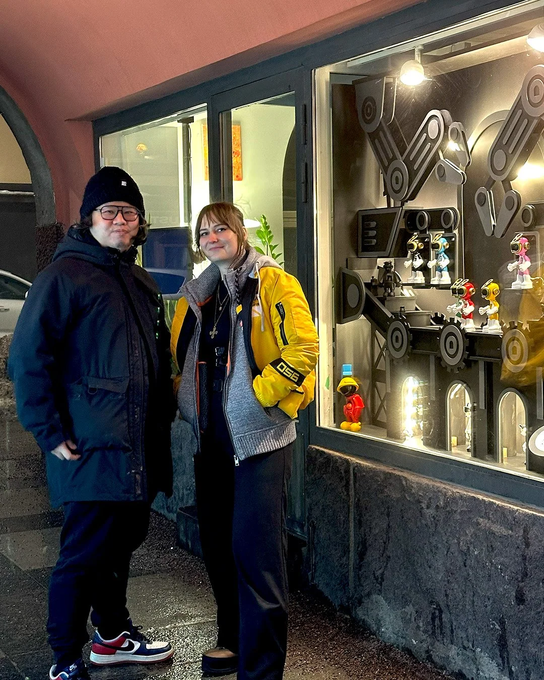 Two people standing outside a store window display featuring various art toys and figures, illuminated with bright lights, at night.