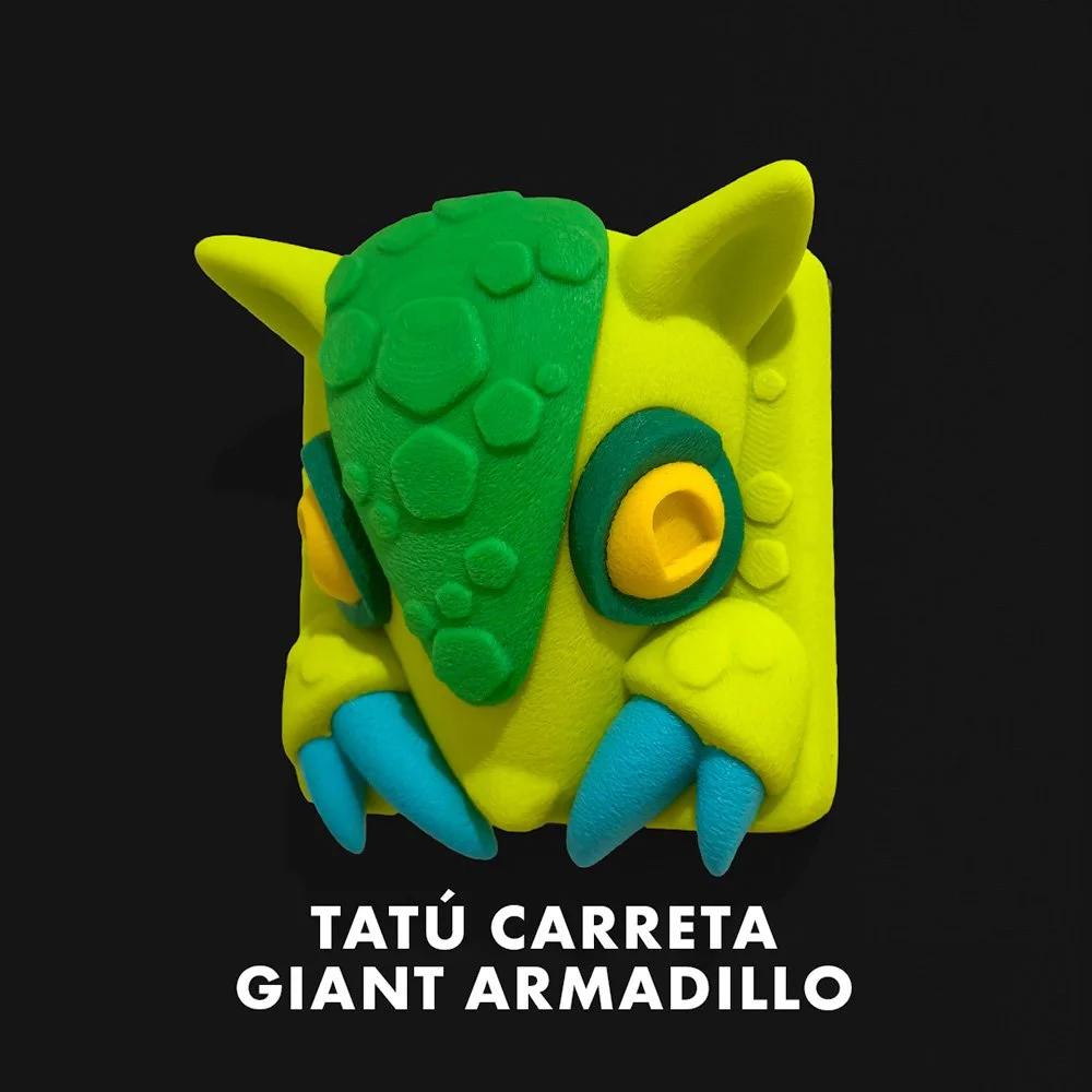 Colorful clay sculpture of a cartoon-style yellow fish with blue fins and green and yellow details, against a black background. Text reads 'Tatú Carreta Giant Armadillo'.