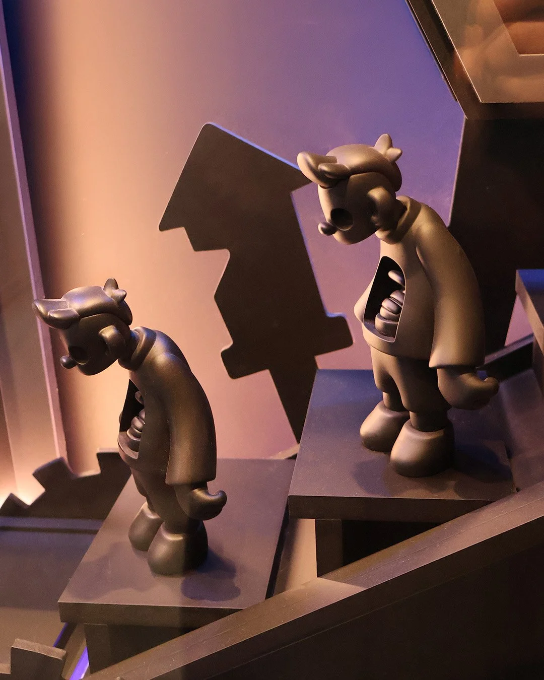Two black joyboy art toy
 with exaggerated features, standing on black platforms with geometric shapes in the background, lit with purple and orange lighting.
