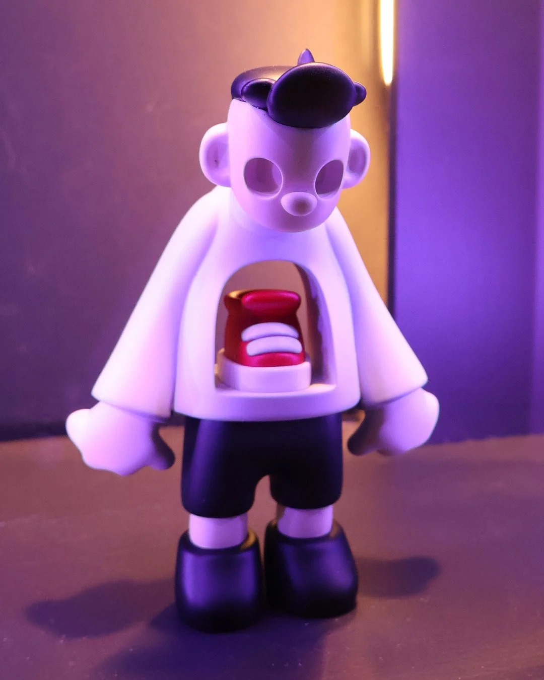 A art toy figure with a cartoonish face, black hair, and large ears. It is holding a red and white object resembling a mouth or device and wears black shorts and boots under a white top. The scene has colorful, purple, and yellow lighting.