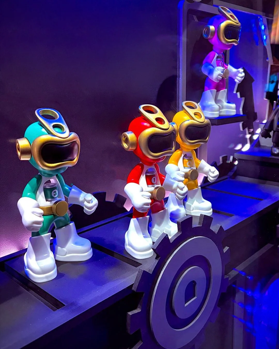 Colorful power ranger art toy figurines in red, yellow, teal, and purple on a black platform with gear-shaped decorations, illuminated by blue and purple lighting.