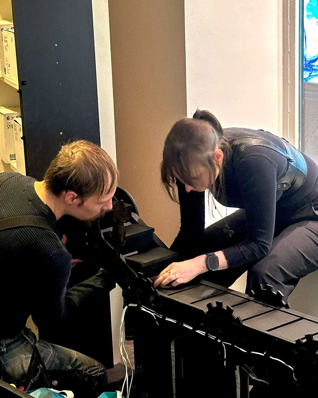 Two individuals working together on a black electronic device, possibly a keyboard, in an indoor setting.