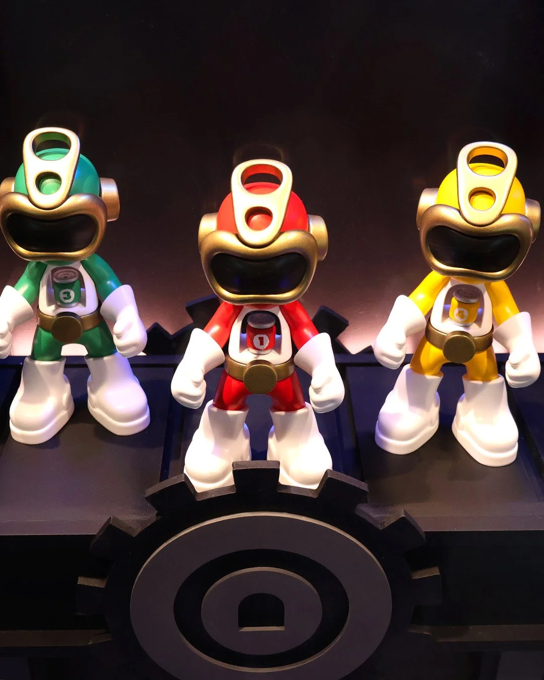 Three statue figures resembling racing cars with exaggerated faces, colored green, red, and yellow, standing on black gear-shaped platform, all with open mouths and white gloves.