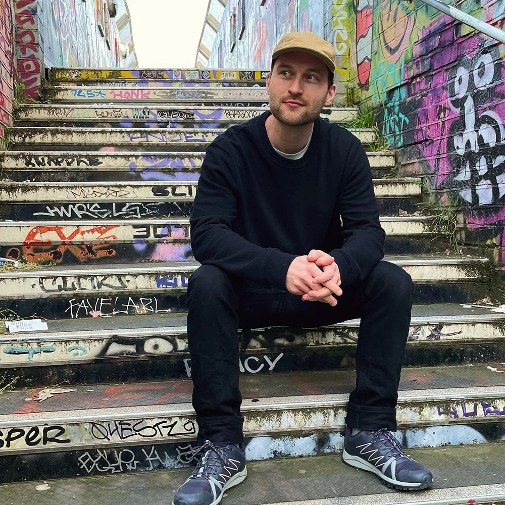 Portrait of a male artist seated on a sticker-covered staircase surrounded by colourful street art.