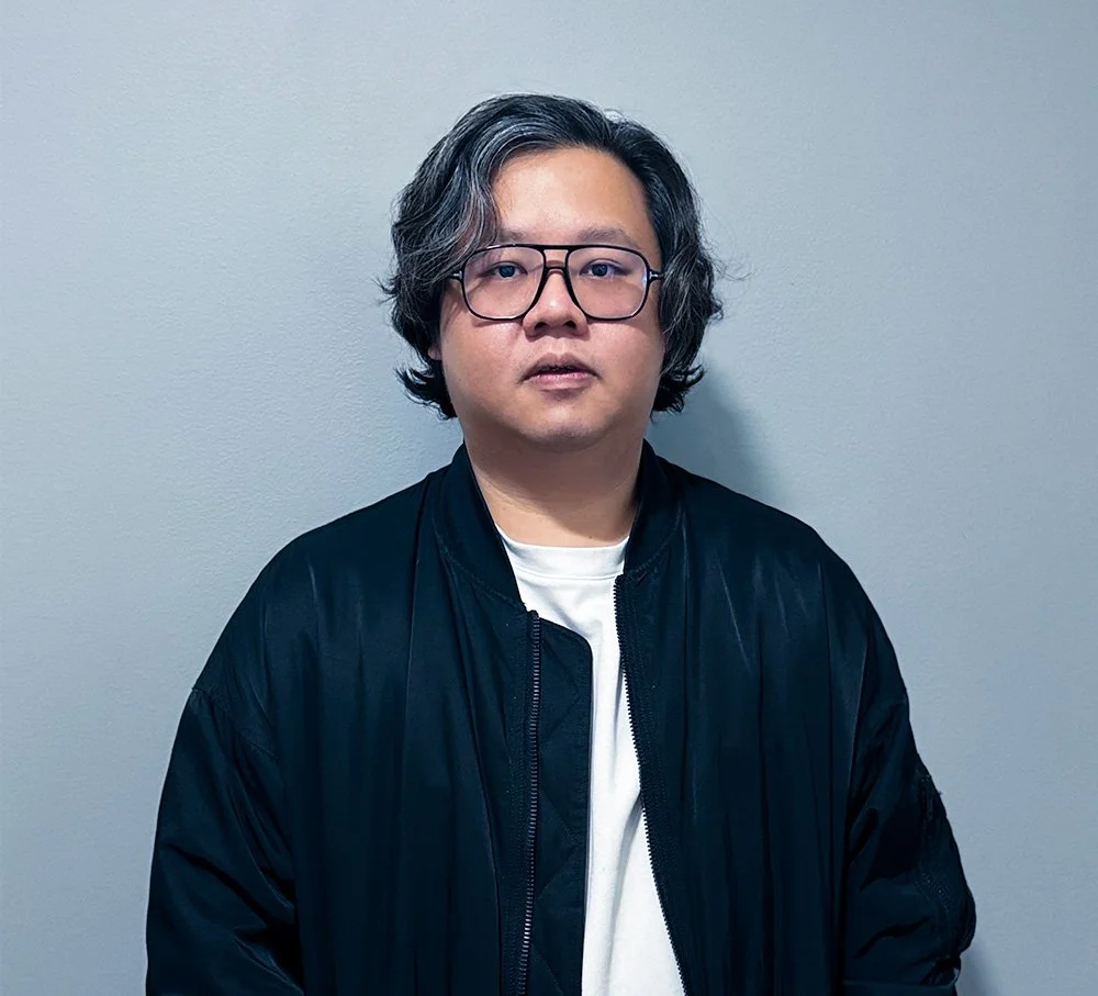 A man with shoulder-length wavy gray hair wearing glasses, a black jacket, and a white shirt standing against a plain gray wall.