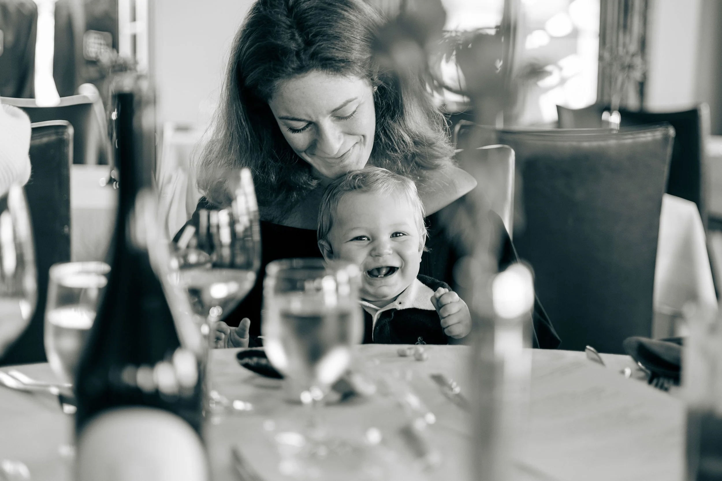 Black and white photo of a smiling woman with a laughing baby at a dining table, surrounded by glasses and a bottle.