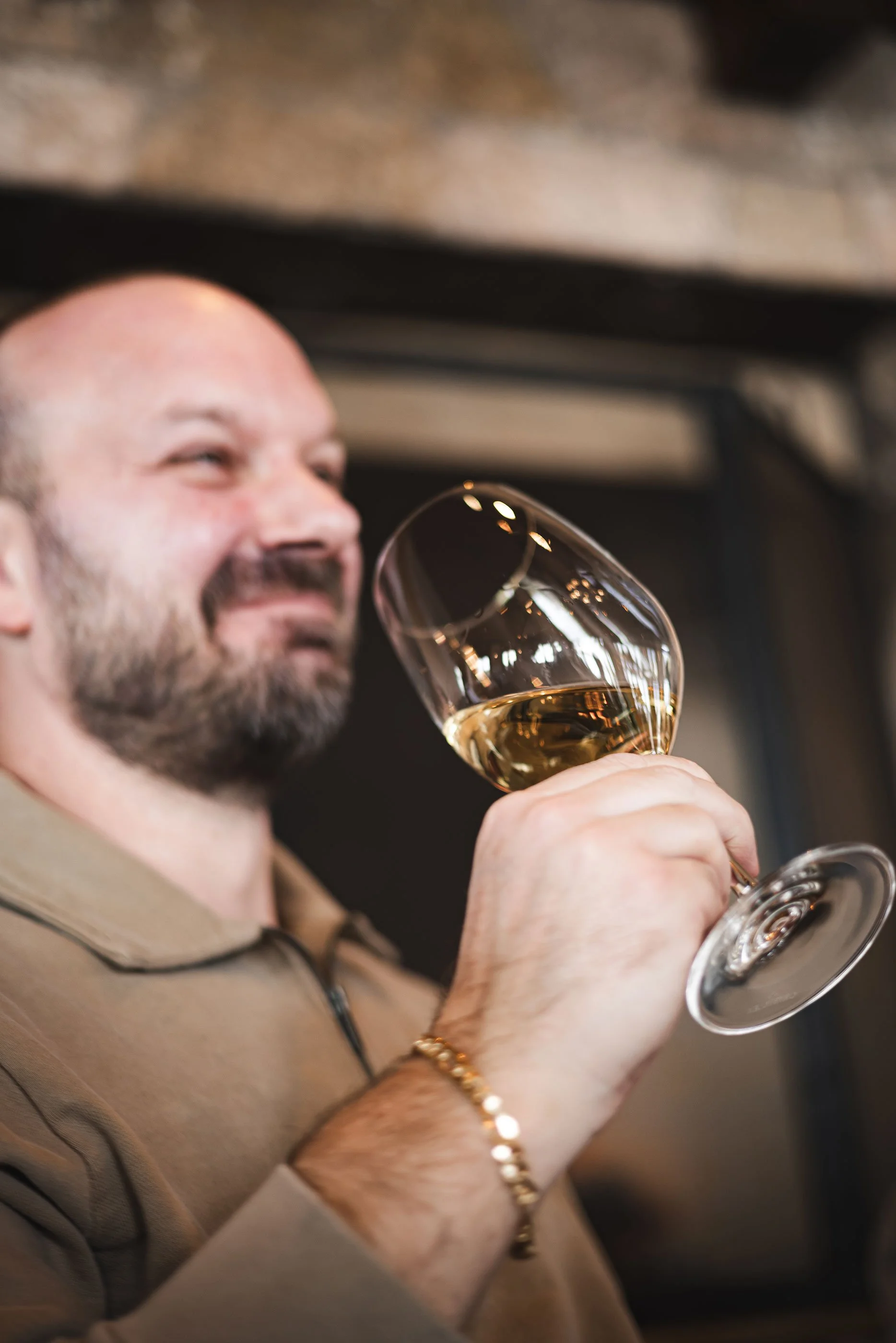 Man with a beard holding a glass of white wine, smiling, wearing a beige shirt and a gold bracelet.