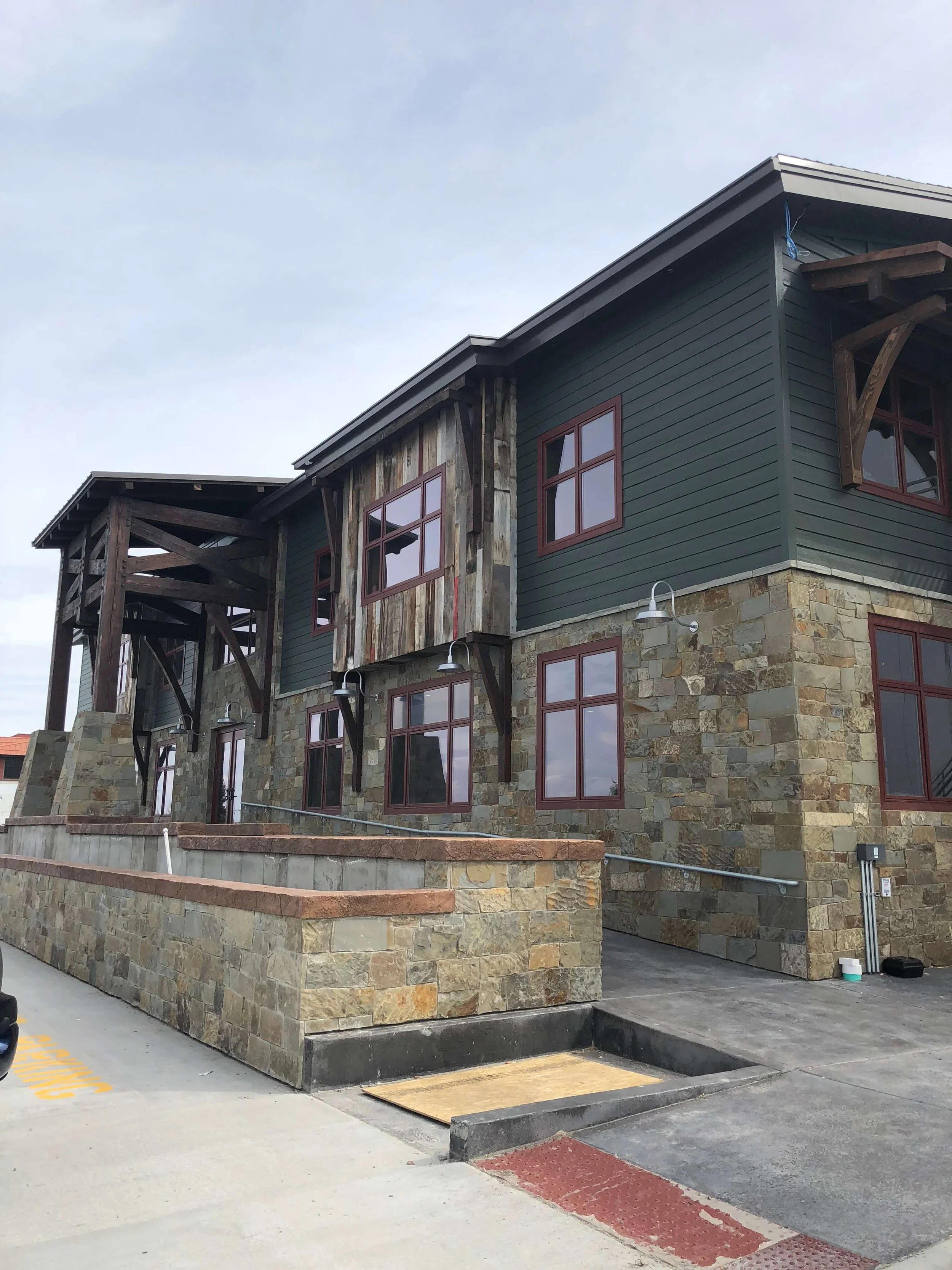 A multi-story commercial business building with a stone and wood exterior, featuring several windows and outdoor lighting fixtures. There is a concrete ramp and parking area in the foreground.