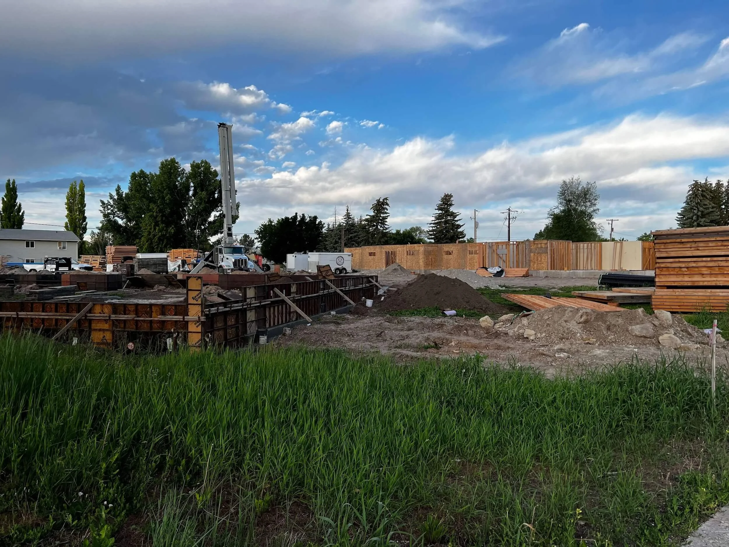 Construction site with wooden framing, dirt piles, construction equipment, and a partly cloudy sky.