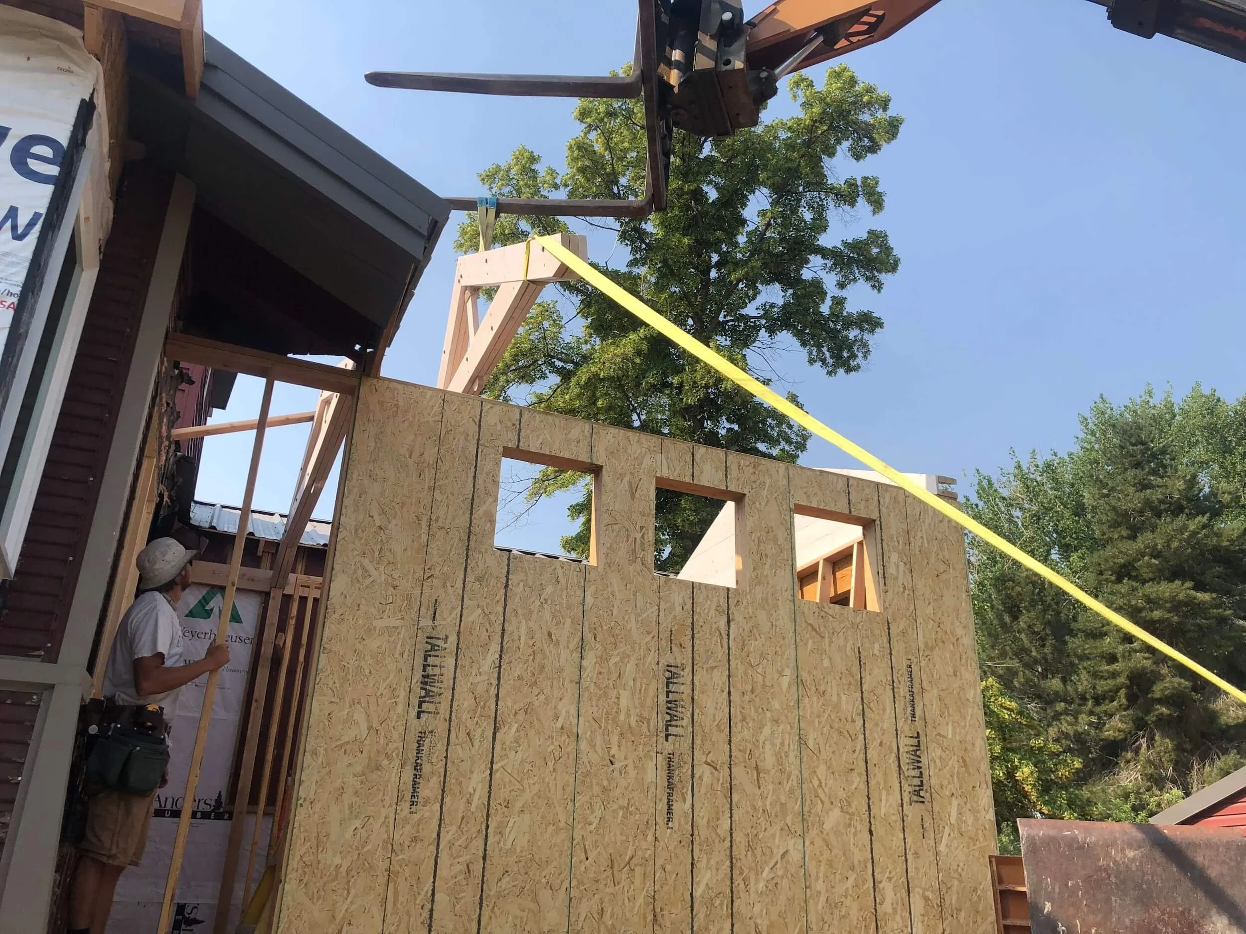 Construction site where wooden wall panels are being installed on a building, with a worker standing below holding a tool, and a yellow measuring tape stretched across the structure, surrounded by trees and blue sky.