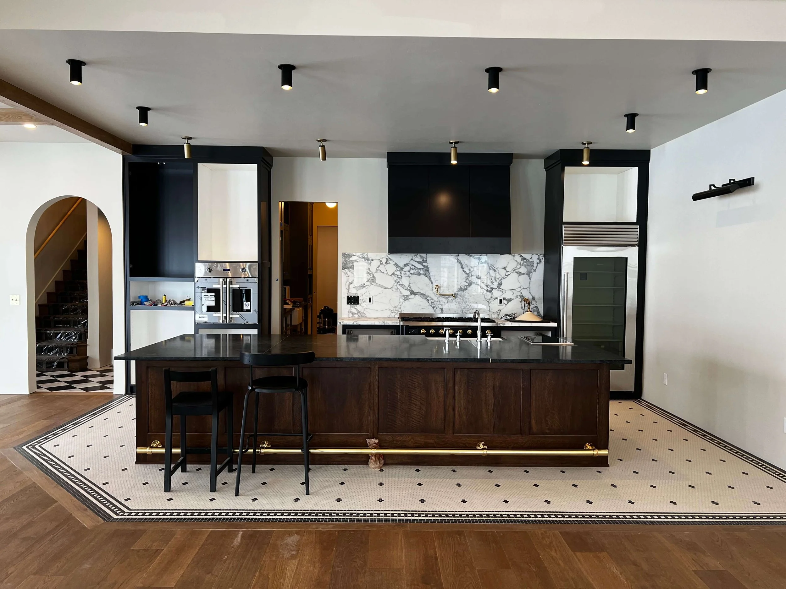 Modern kitchen with dark cabinetry, marble backsplash, and a large island with black countertop and barstool. Hardwood and tiled flooring, small ceiling spotlights, and white walls.