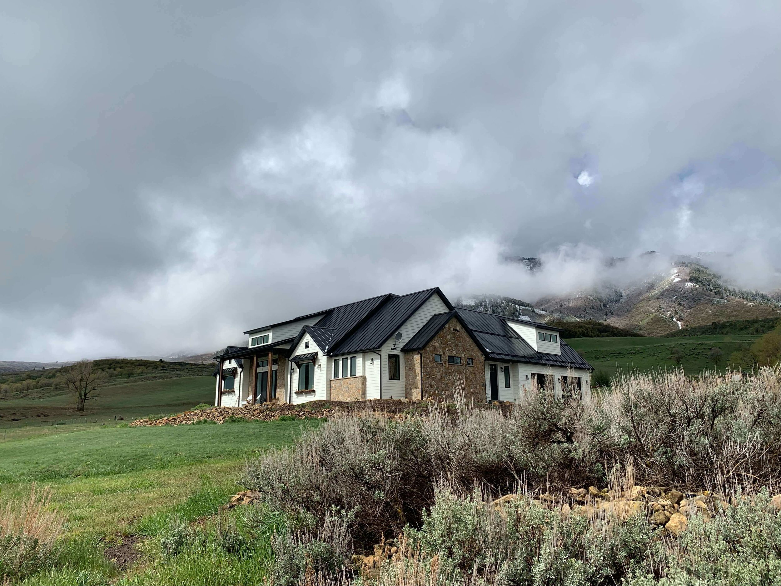 Modern house with stone facade and dark metal roof in a grassy field, surrounded by sagebrush, with mountains and fog in the background.
