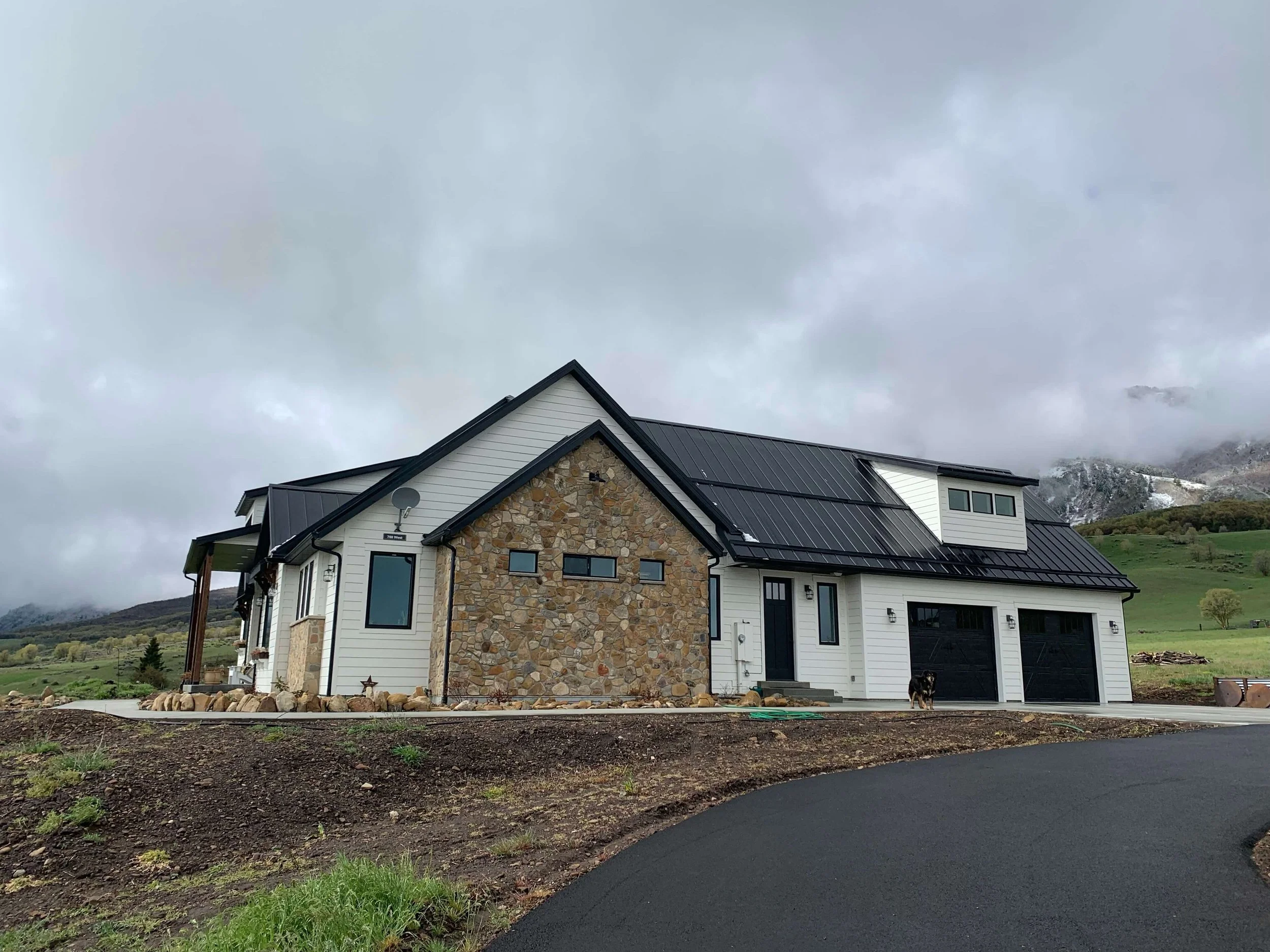 A house with a combination of stone and white siding exterior, a black metal roof, and a small yard with a black dog standing near the garage. The background features cloudy skies and green hills.