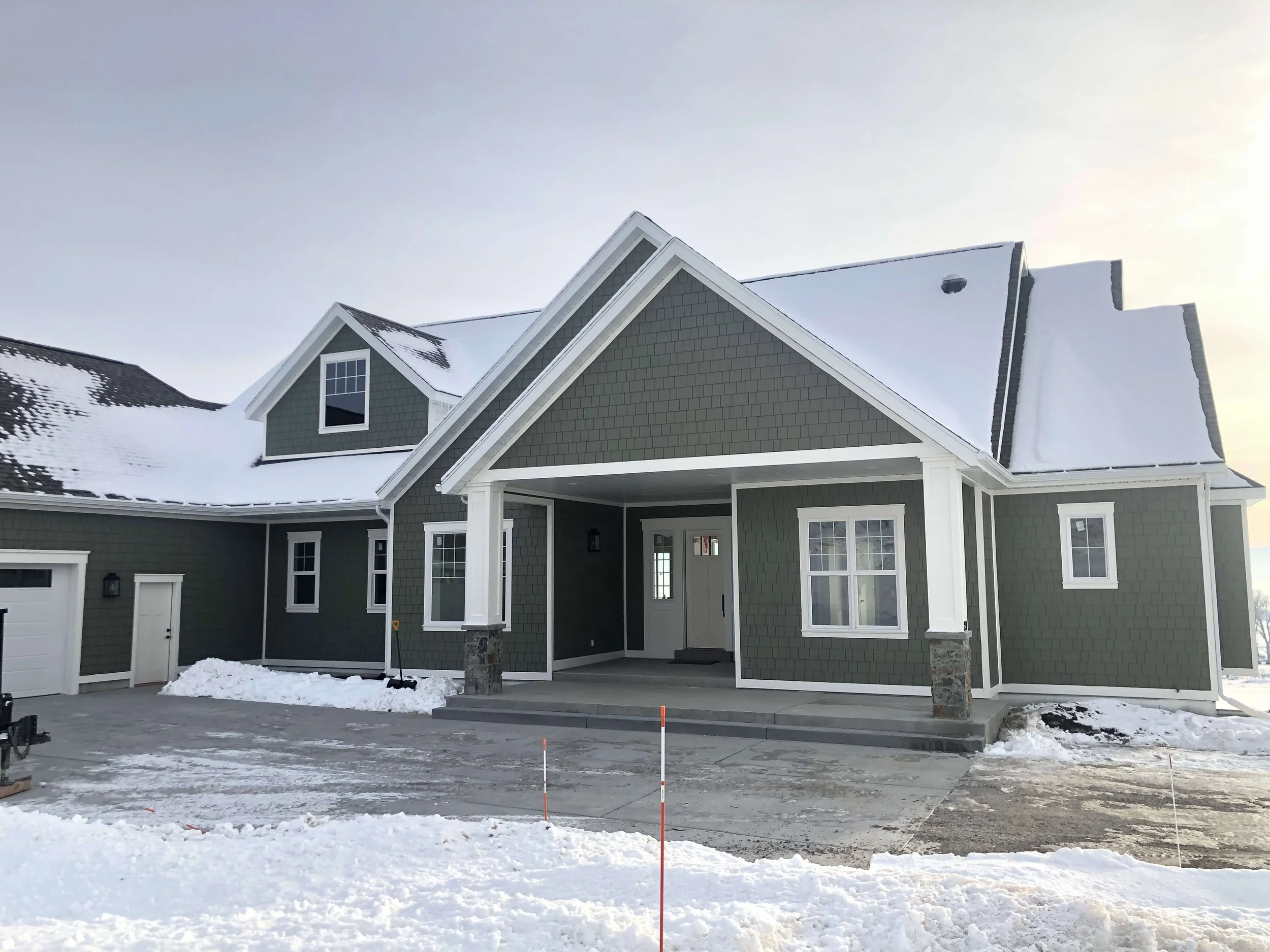 Newly constructed two-story house with green siding, white trim, and a gable roof covered with snow. There is a large front porch, a garage, and a partially snow-covered driveway. The ground and surroundings have snow, with some construction markers visible.