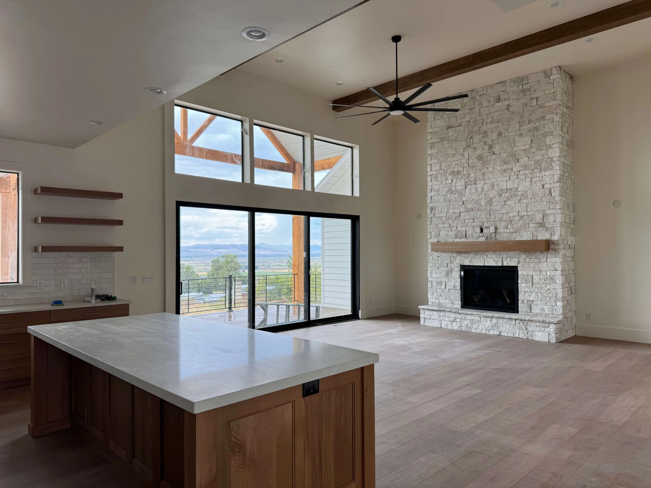 Open-concept living room with large windows, stone fireplace, ceiling fan, and view of outdoor patio and distant landscape.