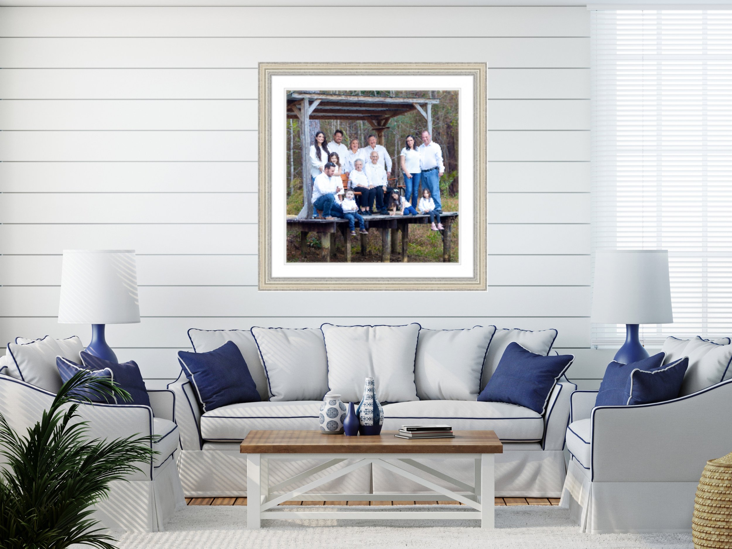 Living room with white and navy striped sofa, navy throw pillows, green plant, wooden coffee table with blue and white vases, large framed family photo on white paneled wall, two white table lamps, window with blinds.