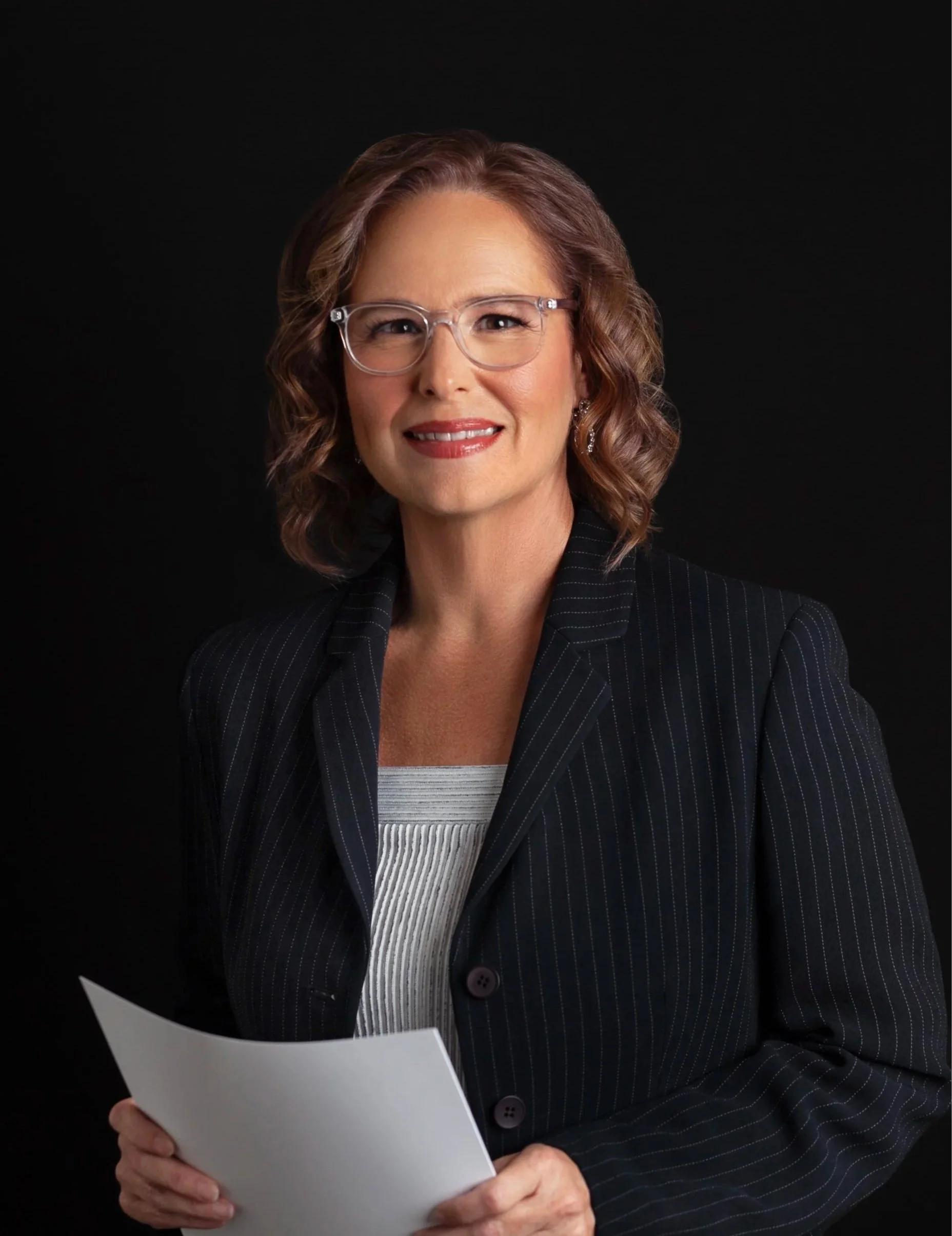 A woman with wavy shoulder-length hair, wearing glasses and a pinstripe blazer, is holding a white piece of paper or document and smiling against a black background.