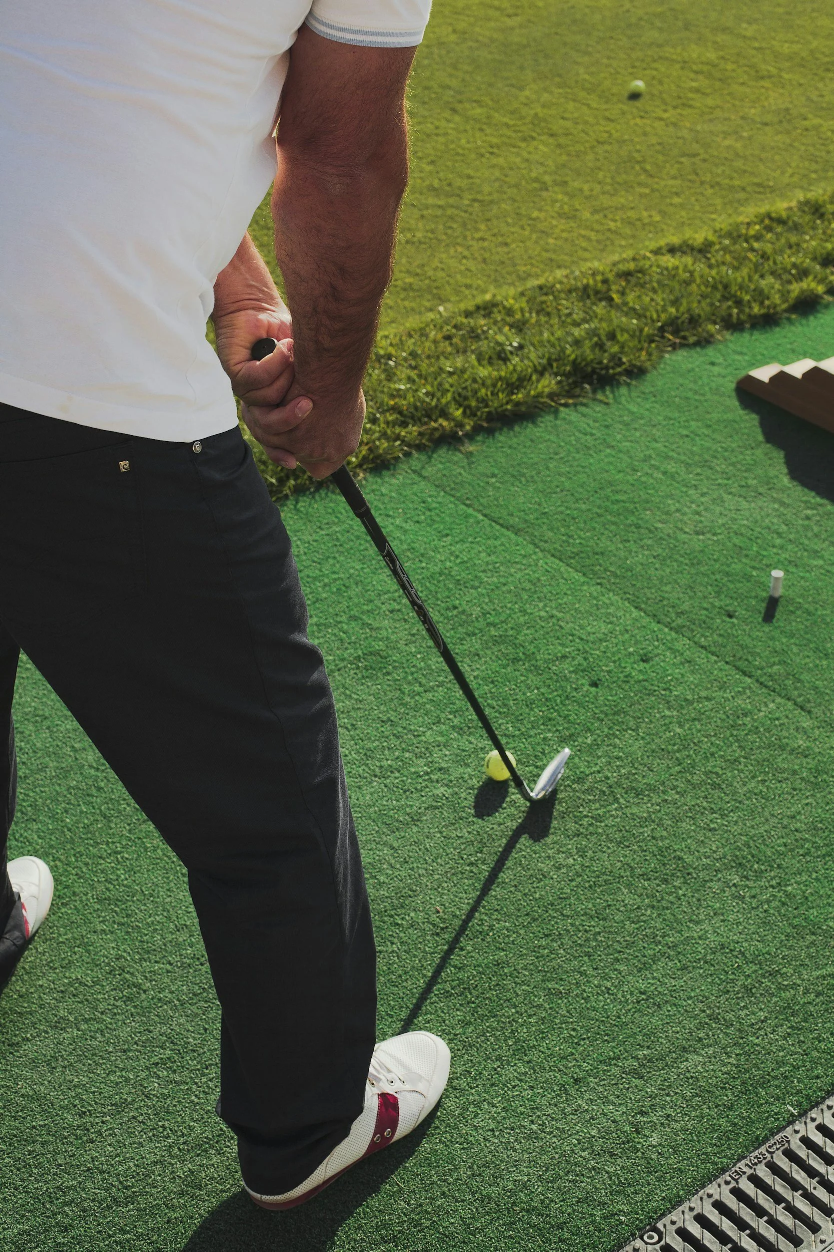 A person preparing to hit a mobile mini golf ball on a putting green, holding a golf club with both hands, wearing a white and blue striped shirt, black pants, and white golf shoes with red accents. 