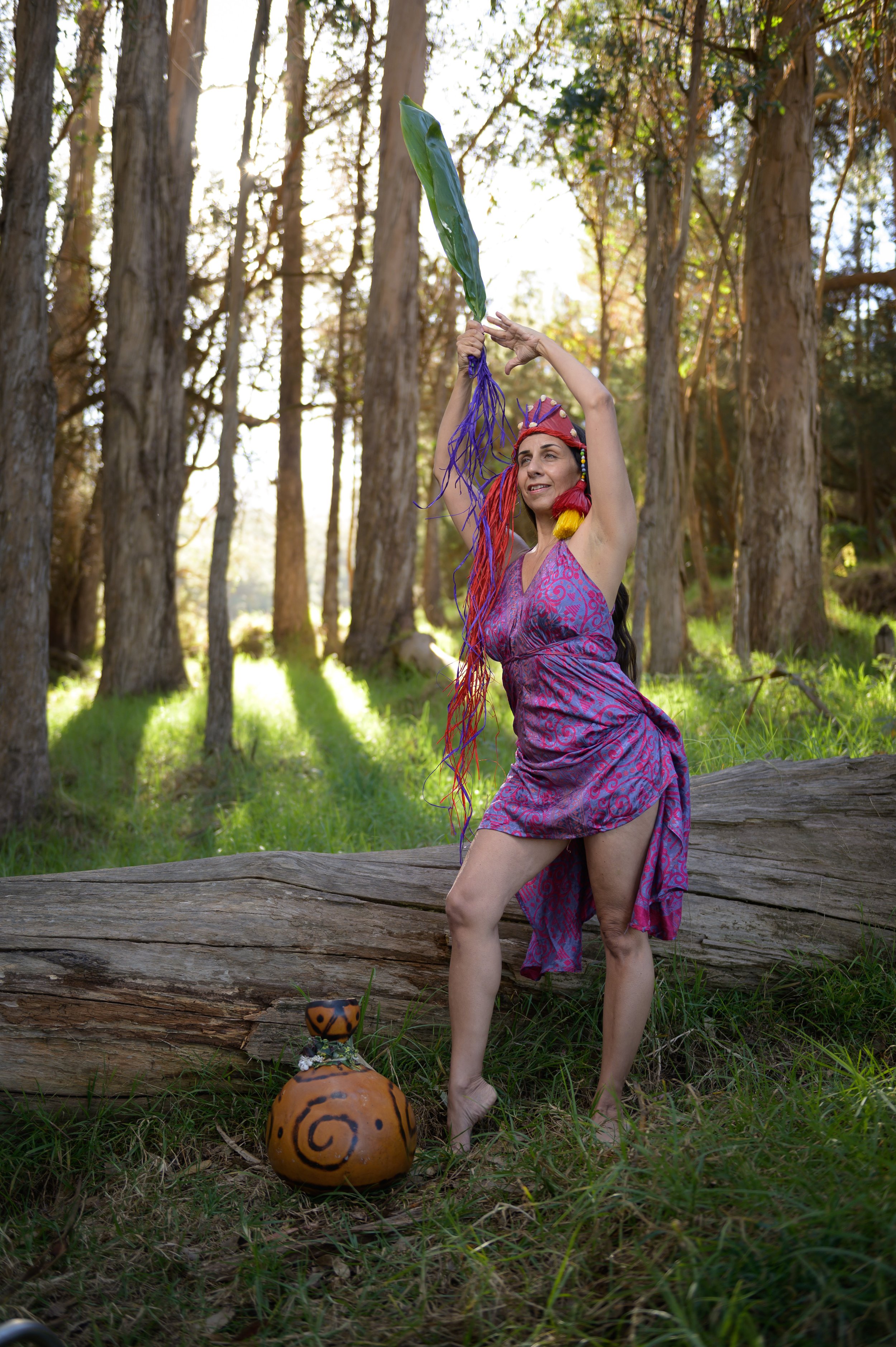 A woman wearing a purple dress with colorful hair accessories holding a large green leaf on a stick in a forest with tall trees and sunlight filtering through the branches. There is a painted ceramic sculpture of a snail on the ground nearby.