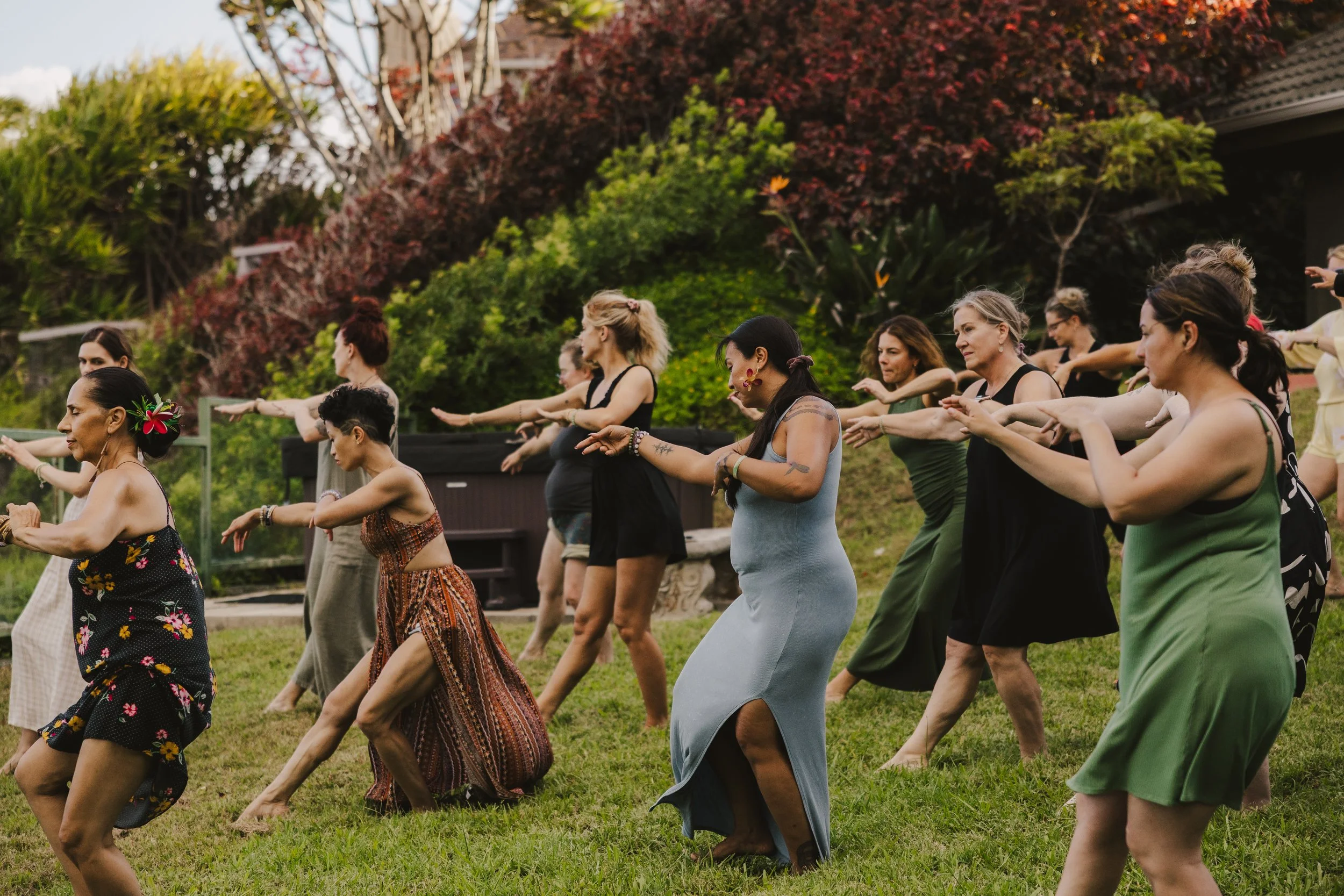 A group of women practicing yoga outdoors on a grassy area with trees and bushes in the background.
