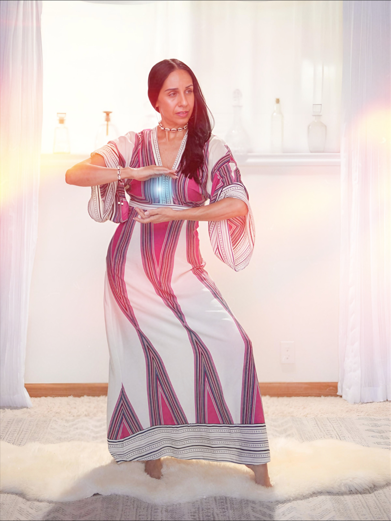 A woman in a colorful striped dress is dancing on a white fluffy rug in a room with light-colored walls and curtains. She is holding a book or tablet, and the room has decorative items on the shelf in the background.